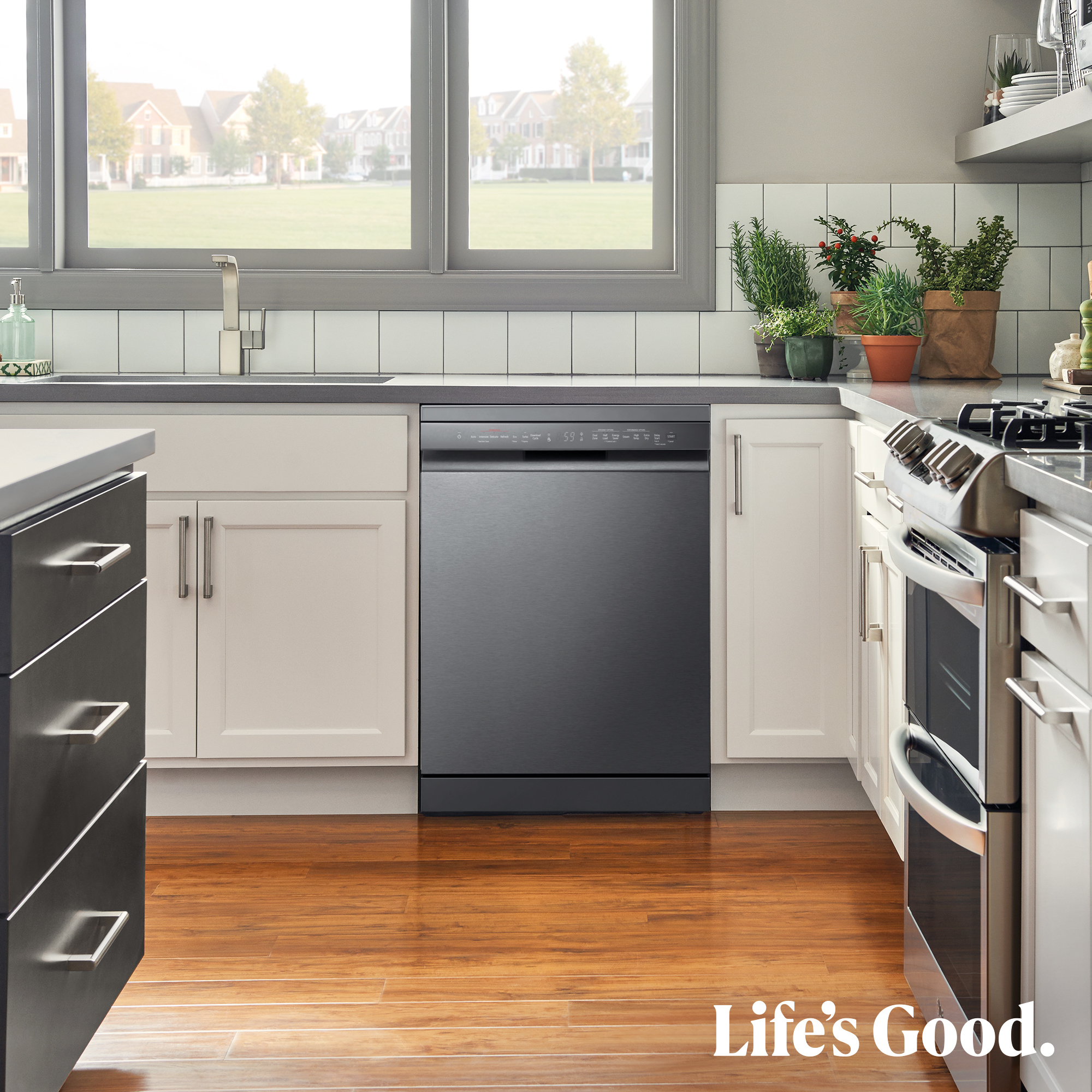 Cocina con lavavajillas negro, armarios blancos, suelo de madera y ventana con vistas a casas. Life's Good está escrito en el suelo.