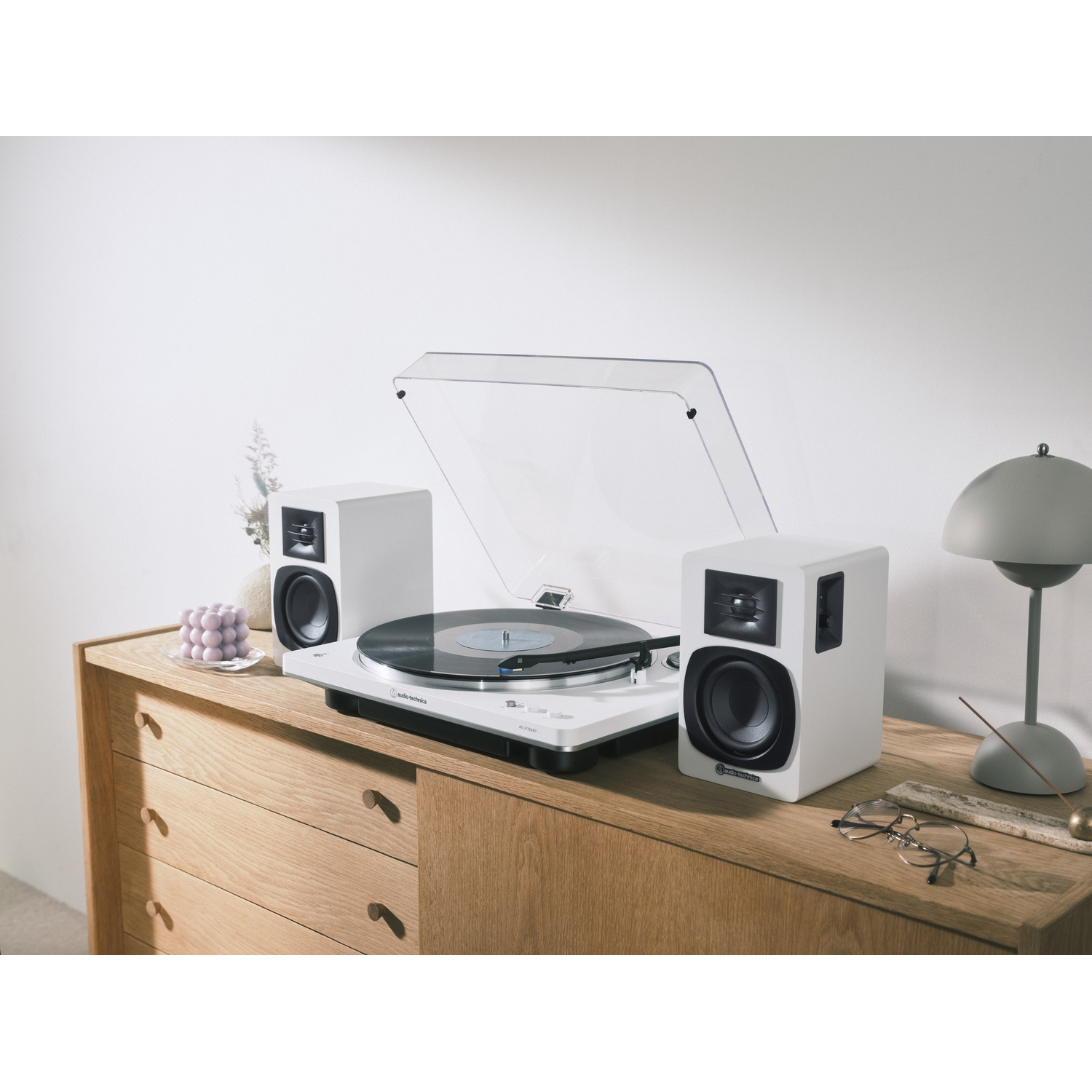 Un giradischi bianco con altoparlanti è appoggiato su un mobile di legno. Sono presenti anche una lampada, occhiali e decorazioni.