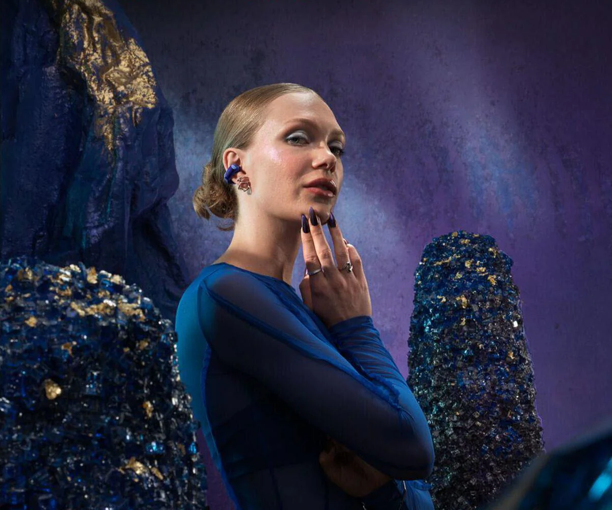 Eine Frau in blauem Kleid posiert, Hand am Gesicht, vor einem Hintergrund aus dunkelblauen Felsformationen. Sie trägt Ohrringe und hat lange Nägel.