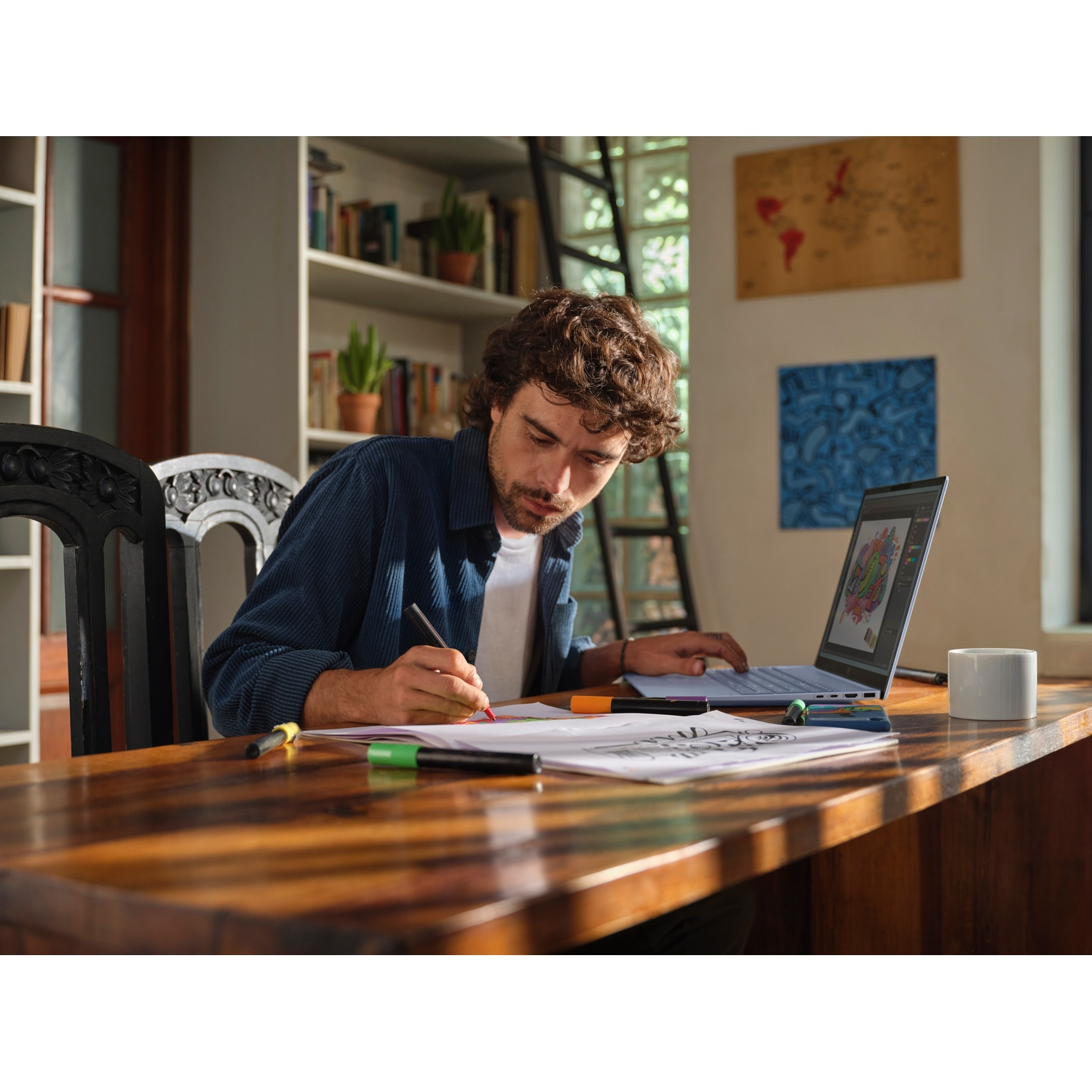 Uomo con camicia blu che disegna su carta. Laptop con design colorato, pennarelli e una tazza sul tavolo. Librerie sullo sfondo.