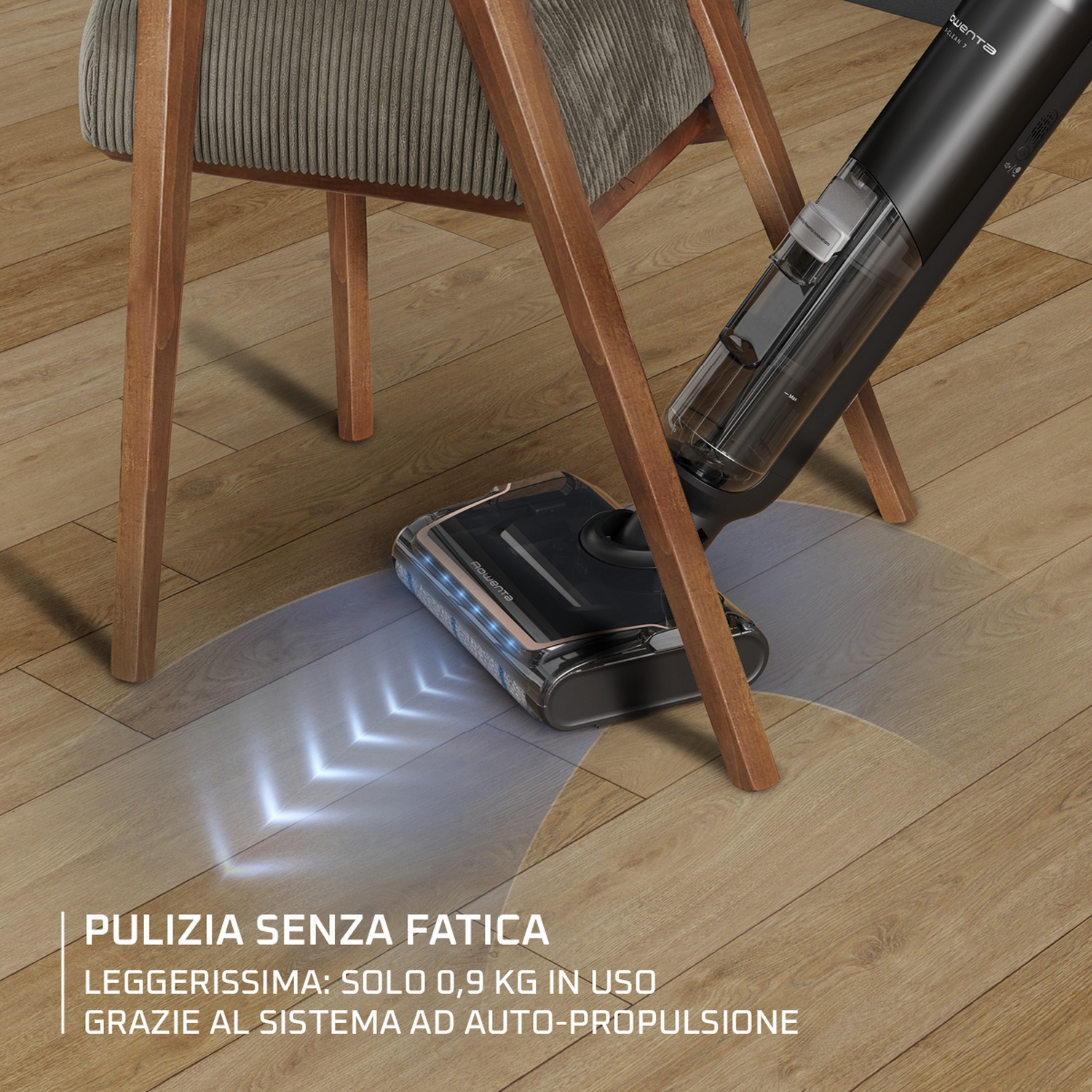 Un aspirapolvere nero pulisce un pavimento in legno. C'è una sedia dietro. L'aspirapolvere è vicino alle gambe della sedia.