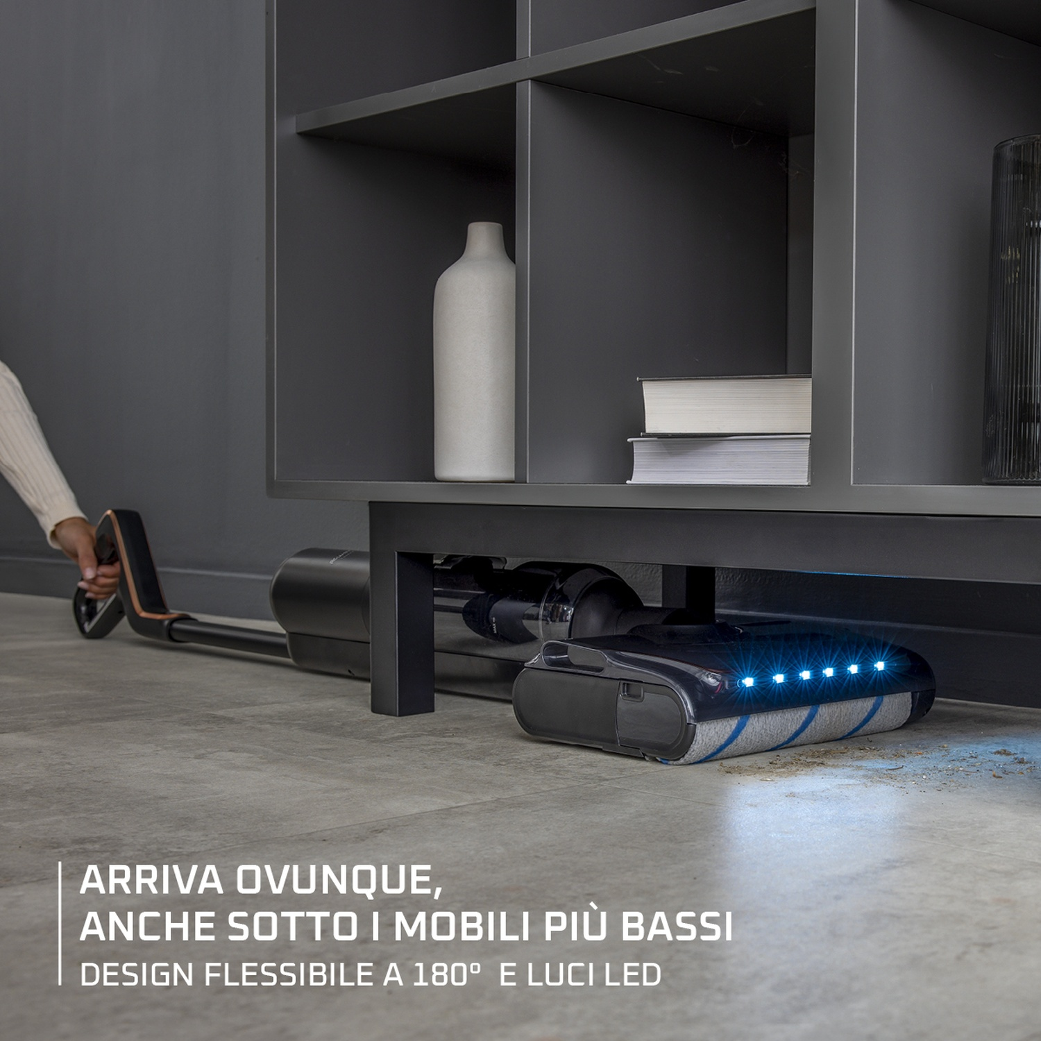 Una persona usa un aspirapolvere sotto un mobile. L'aspirapolvere ha luci a LED. Il mobile ha decorazioni bianche e libri.