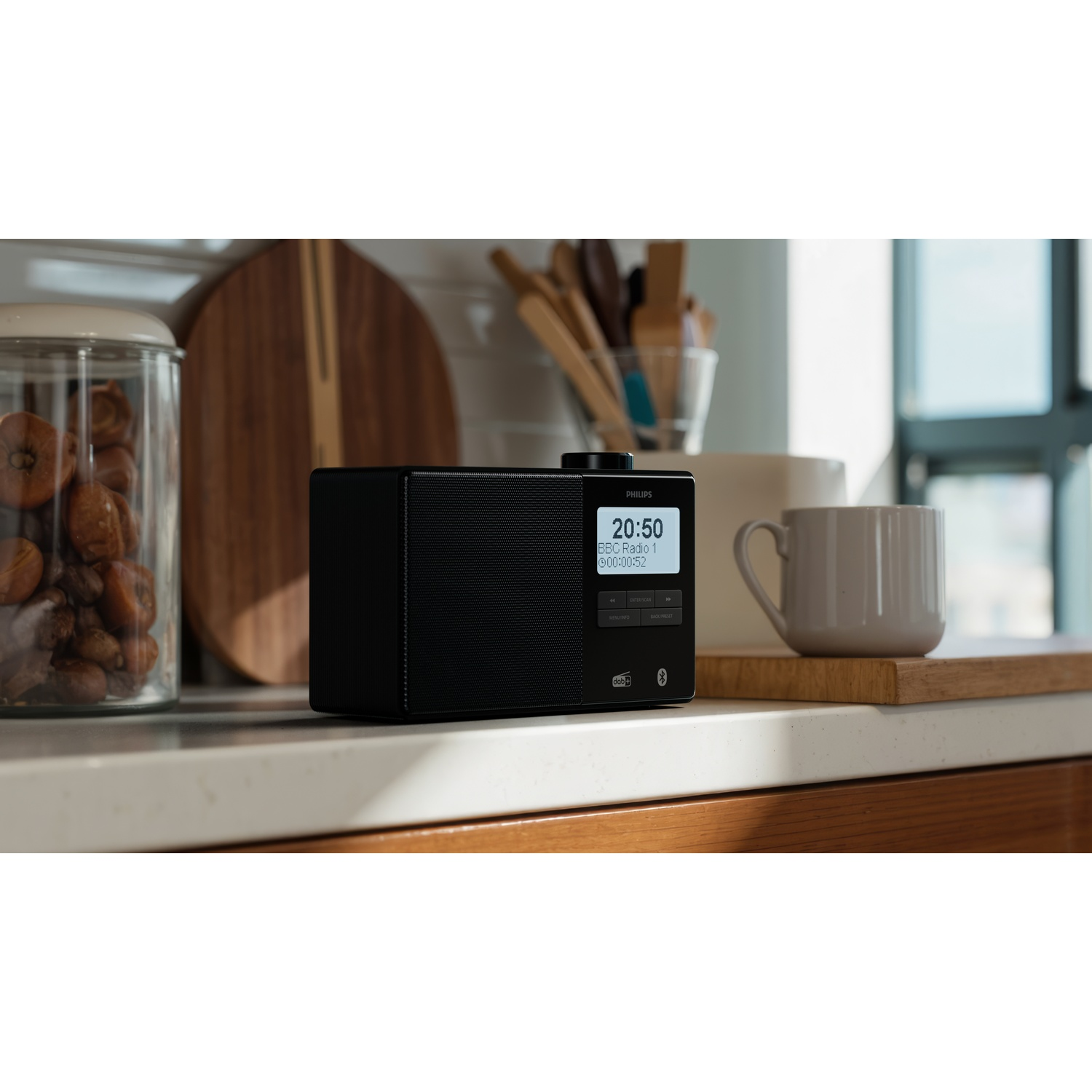 Una radio Philips nera su un bancone. Tazza e assi di legno vicine. Un barattolo con cibo e una finestra sullo sfondo.