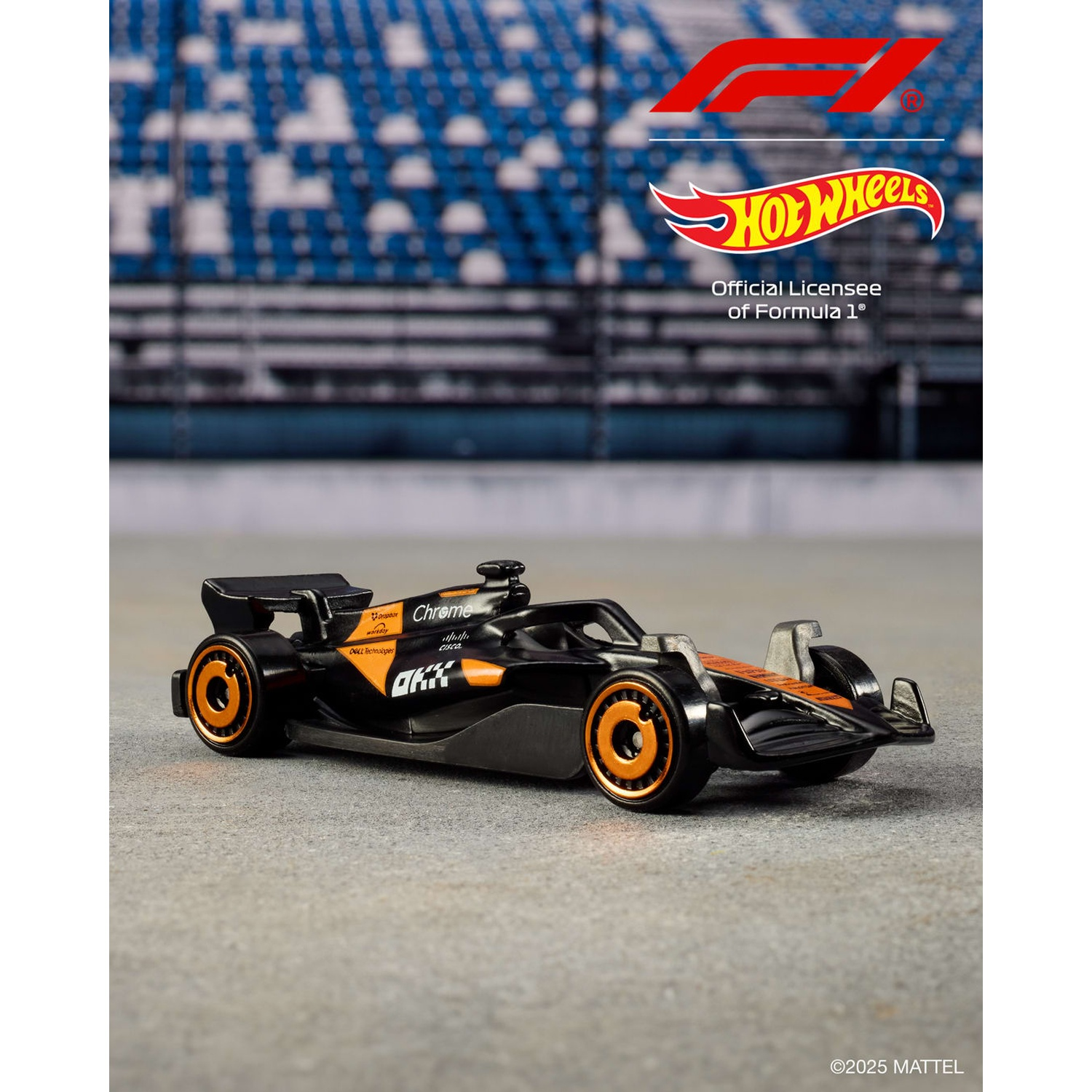 Un'auto di Formula 1 Hot Wheels nera e arancione su una superficie di cemento. Lo sfondo presenta sedili dello stadio e loghi.