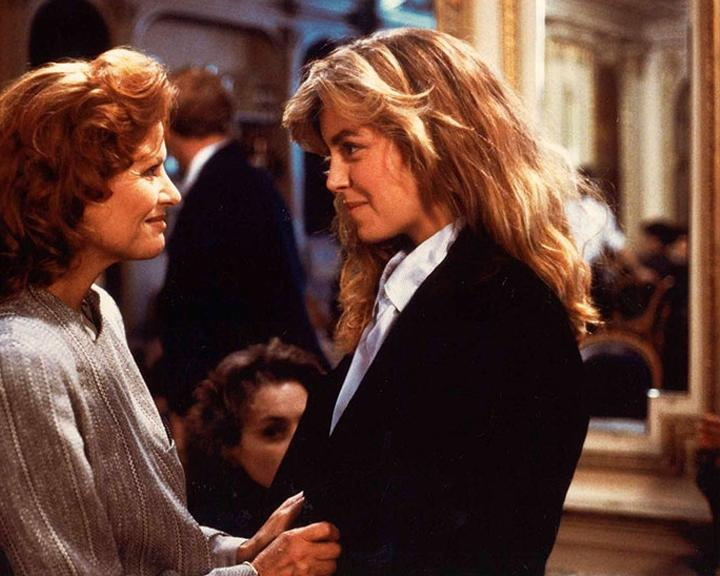 Zwei Frauen stehen sich gegenüber, eine in einem Blazer, die andere in einem Pullover. Andere sind im Hintergrund verschwommen. Beide Frauen lächeln.