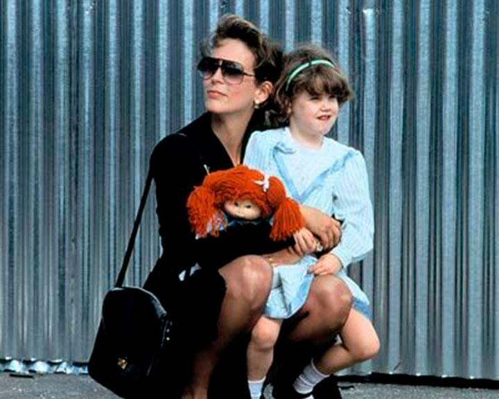 Eine Frau mit Sonnenbrille und ein Mädchen hocken zusammen. Die Frau hält eine Puppe. Im Hintergrund ist eine Metallwand.