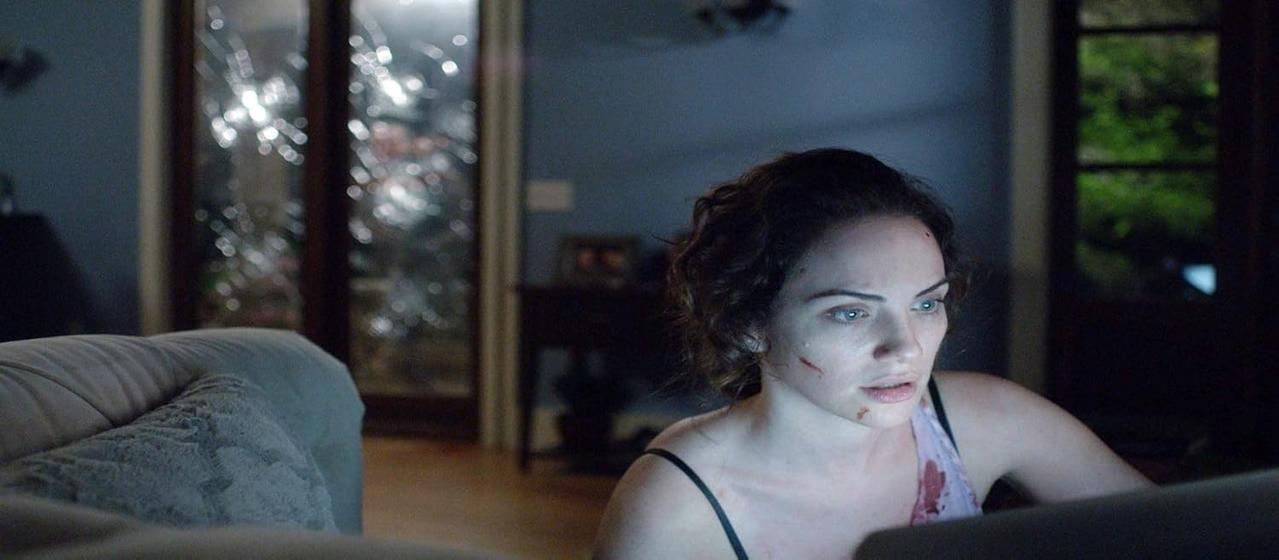 Eine Frau mit Blut im Gesicht blickt auf einen Bildschirm. Eine Couch ist im Vordergrund, ein Fenster im Hintergrund.