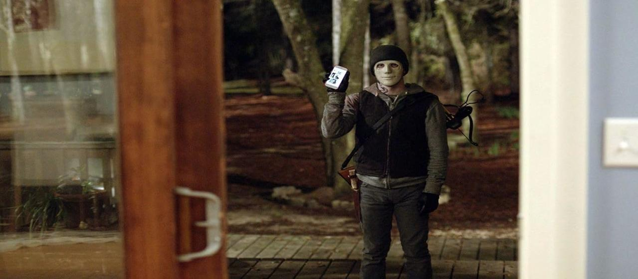 Eine Person mit Maske hält ein Telefon und steht vor einer Tür. Eine Armbrust hängt auf dem Rücken. Im Hintergrund sind Bäume.