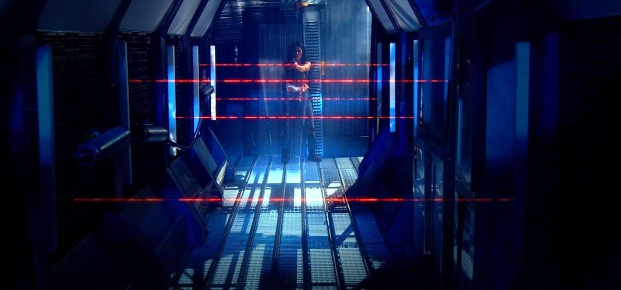Eine Person steht in einem futuristischen Flur. Rote Laserlinien kreuzen den Flur. Die Szene ist dunkel mit blauer Beleuchtung.