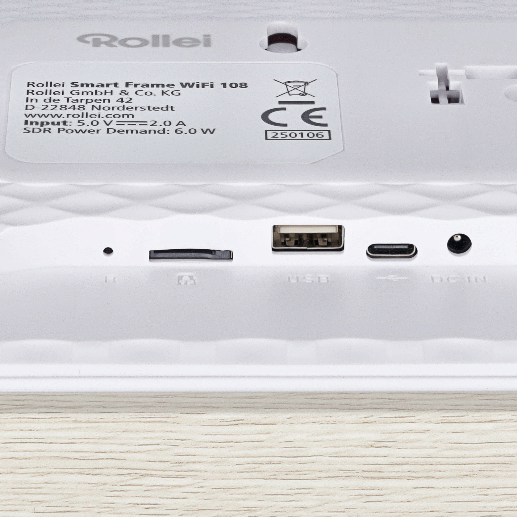 Weißes Gerät mit Anschlüssen, USB und DC IN. Text enthält 'Rollei Smart Frame'. Holzfußboden unten.