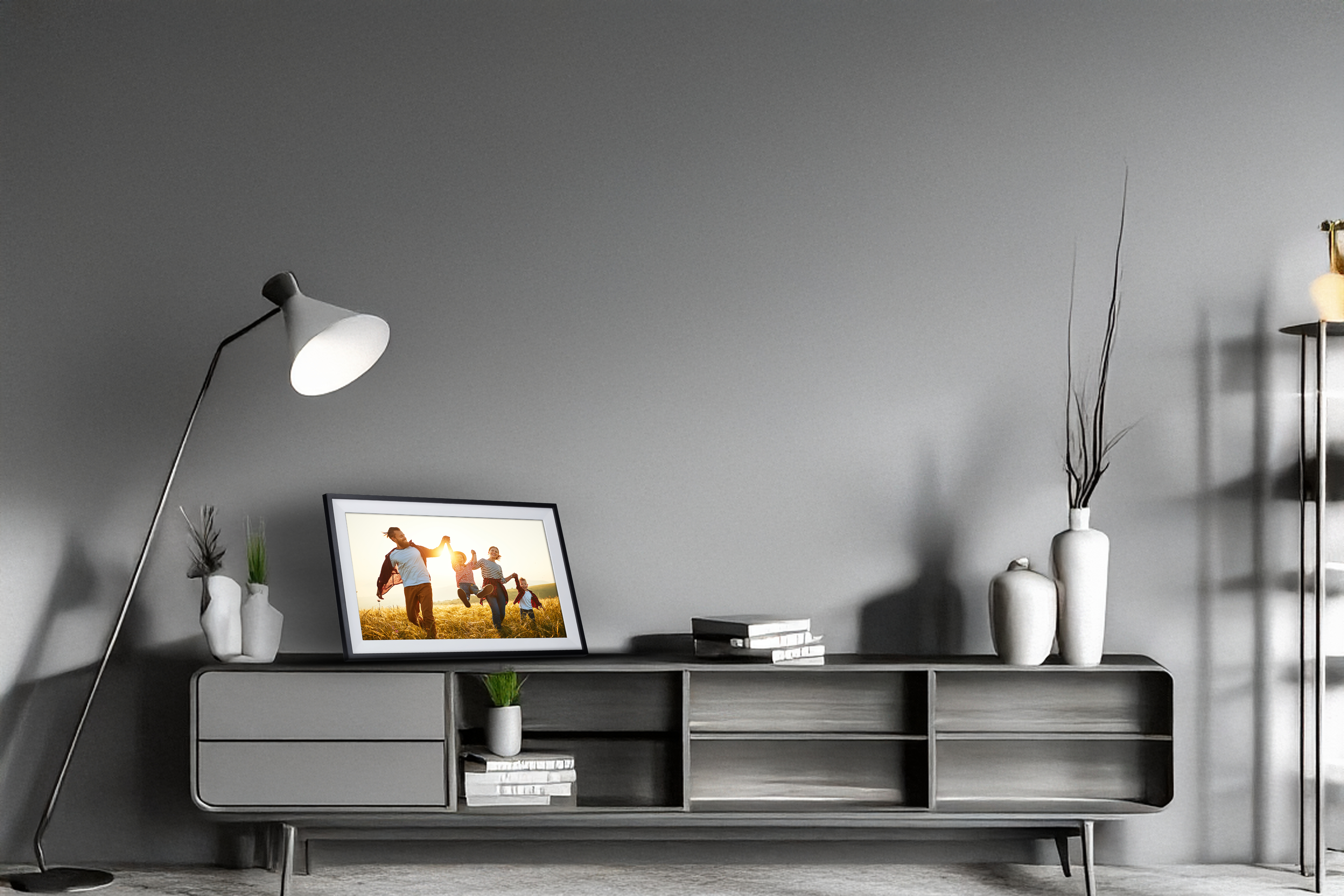 Grauer Schrank mit Deko, Lampe und einem Foto einer Familie. Graue Wand. Die Lampe hat einen weißen Schirm.