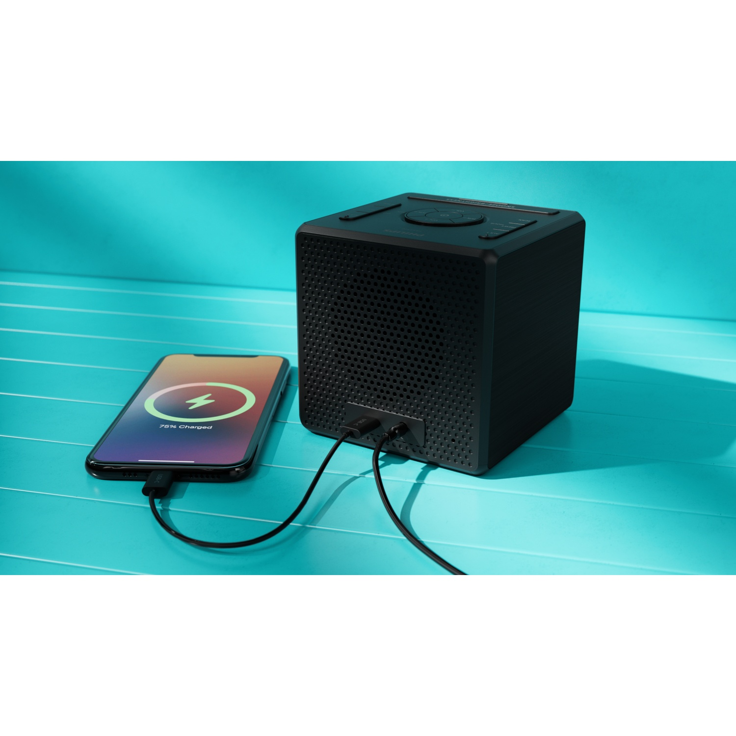 Un altoparlante a cubo nero accanto a un telefono in carica. Il telefono mostra il 75% di carica. Entrambi sono su una superficie turchese.