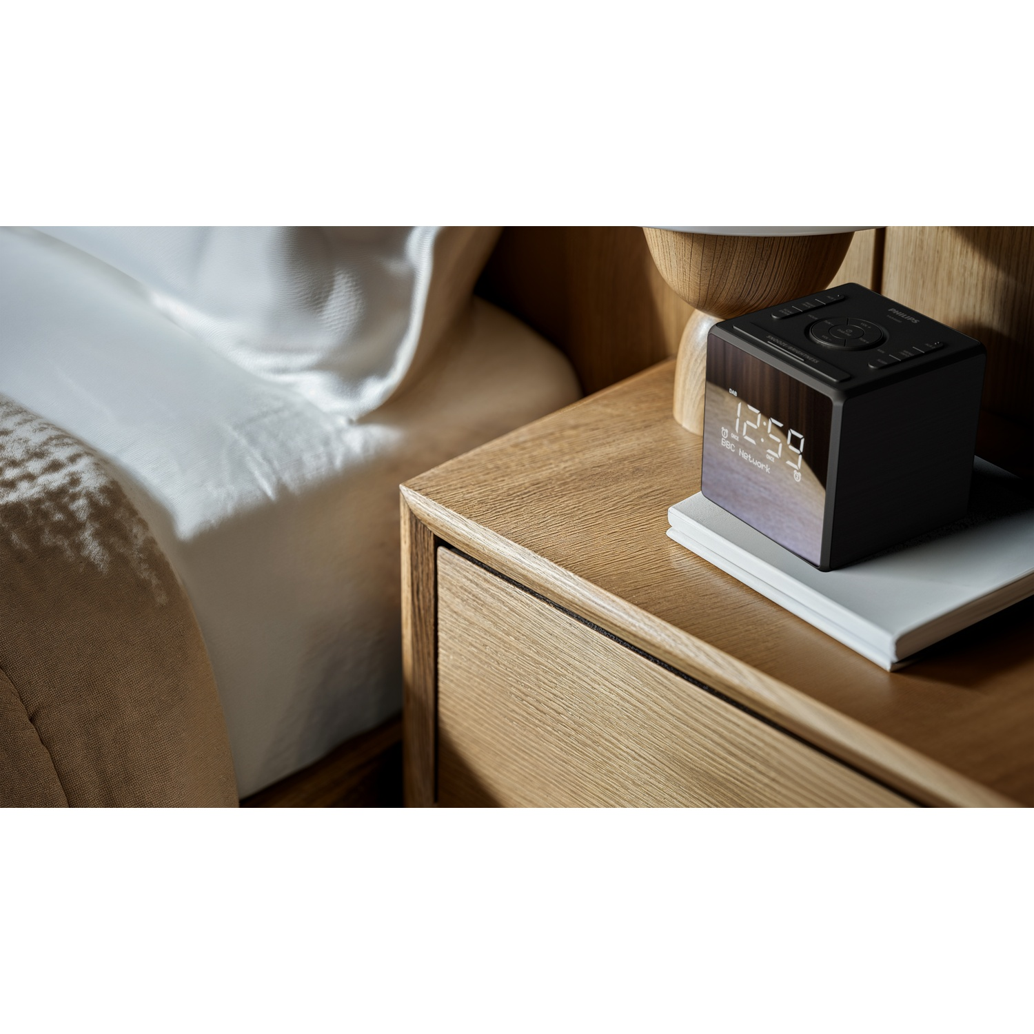 Una sveglia digitale nera che mostra le 12:59 su un comodino di legno, accanto a un letto con lenzuola bianche.