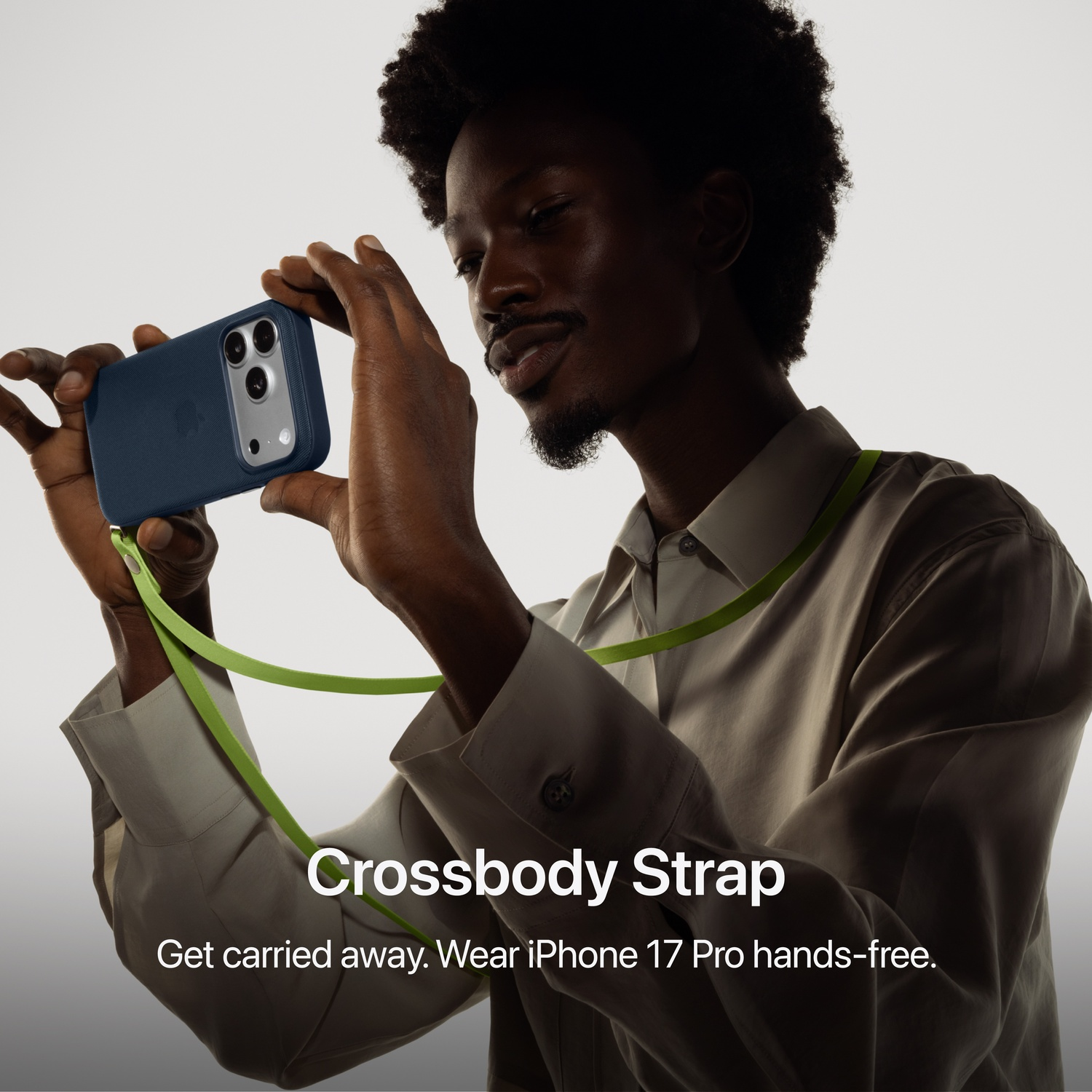Homme tenant un iPhone bleu marine avec une sangle verte. Le téléphone a plusieurs caméras. Texte : Crossbody Strap. Get carried away.