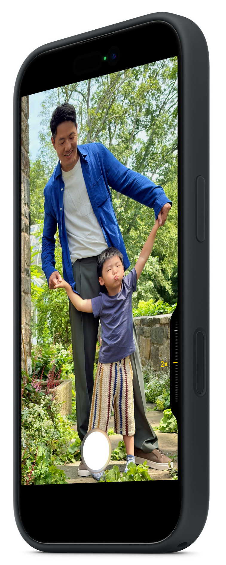 Mężczyzna w niebieskiej koszuli trzyma ręce chłopca, który robi minę. Są na zewnątrz, otoczeni zielonymi roślinami i drzewami.