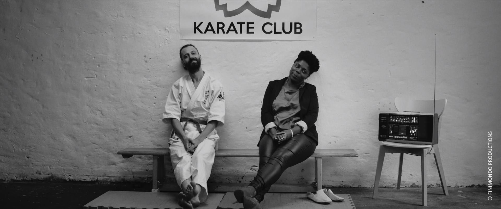 Zwei Personen sitzen auf einer Bank vor einer Wand mit einem Karate-Club-Schild. Auf einem Stuhl steht ein Radio. Schwarz-Weiß-Foto.
