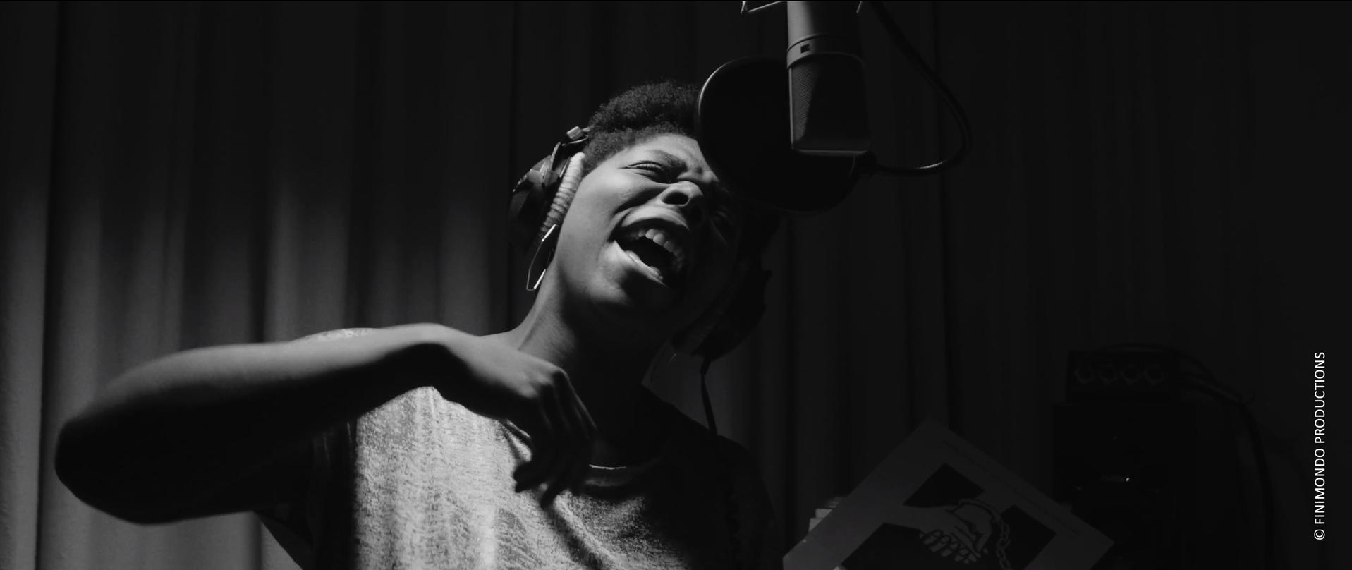 Eine Person singt in einem Studio in ein Mikrofon. Sie tragen Kopfhörer. Ein Papier wird gehalten.