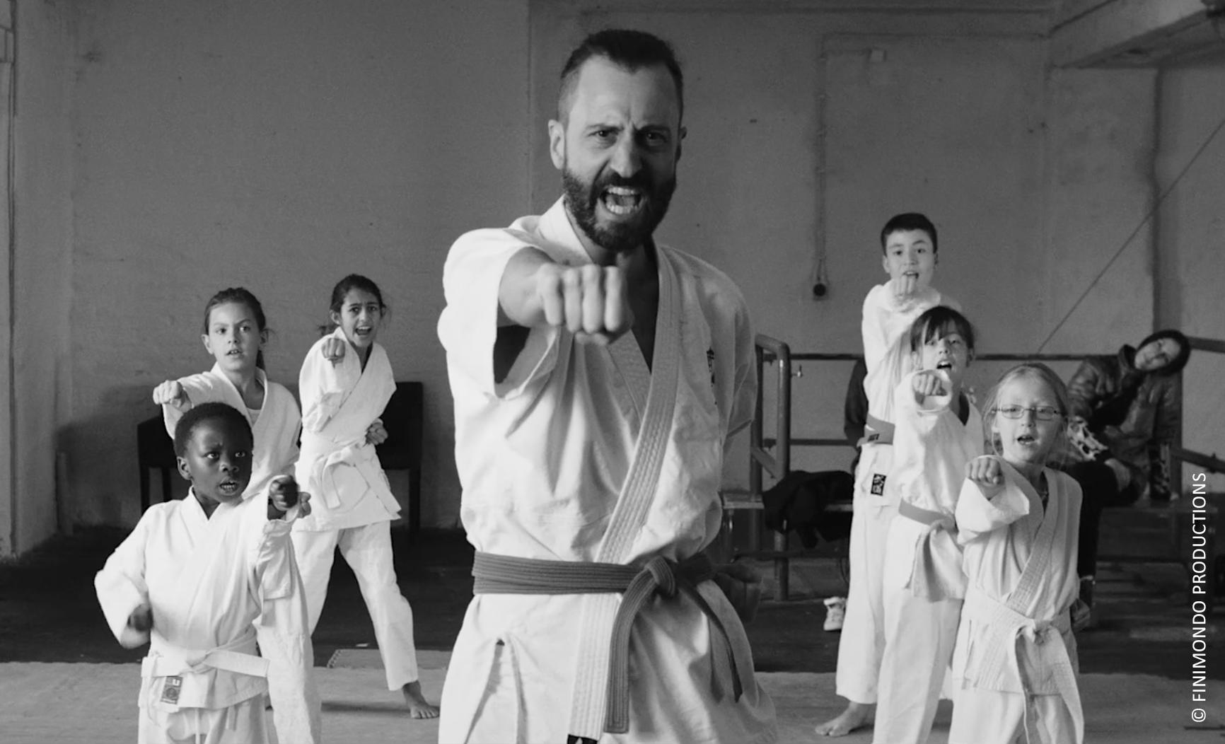Eine Gruppe von Karate-Praktizierenden, darunter ein Mann und Kinder, schlagen nach vorne. Die Kulisse ist eine Trainingshalle, in Schwarzweiß.
