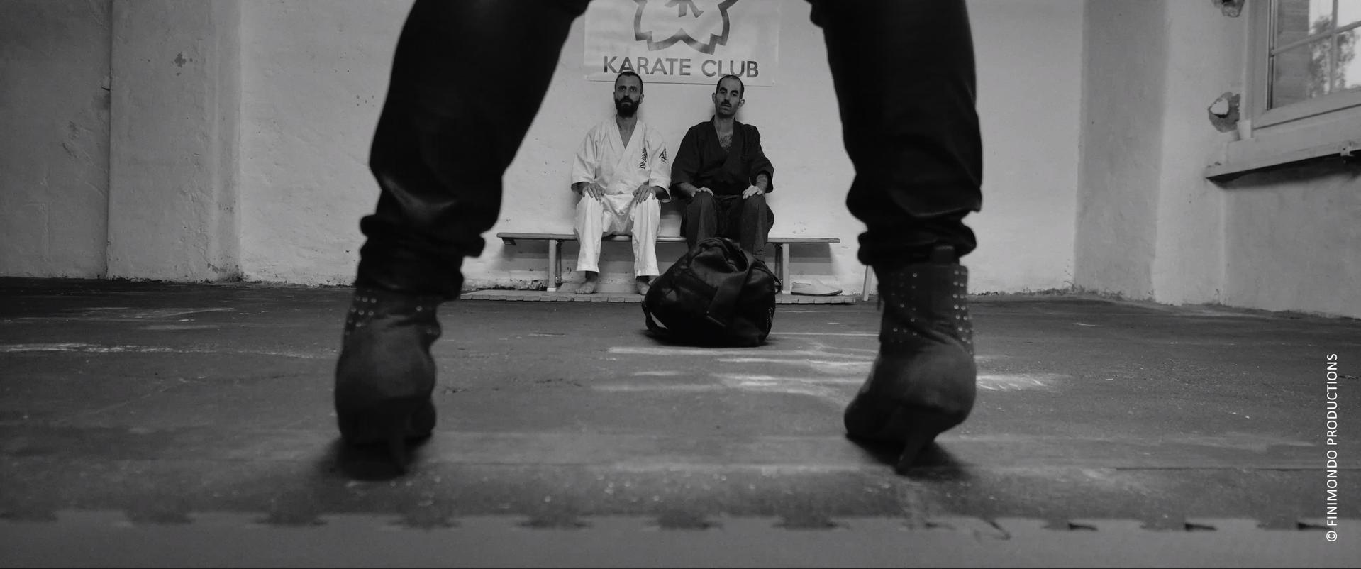 Zwei Männer im Karate-Club, einer in weißer Uniform, der andere in schwarz. Eine Tasche liegt auf dem Boden. Vordergrund: Beine und Stiefel.