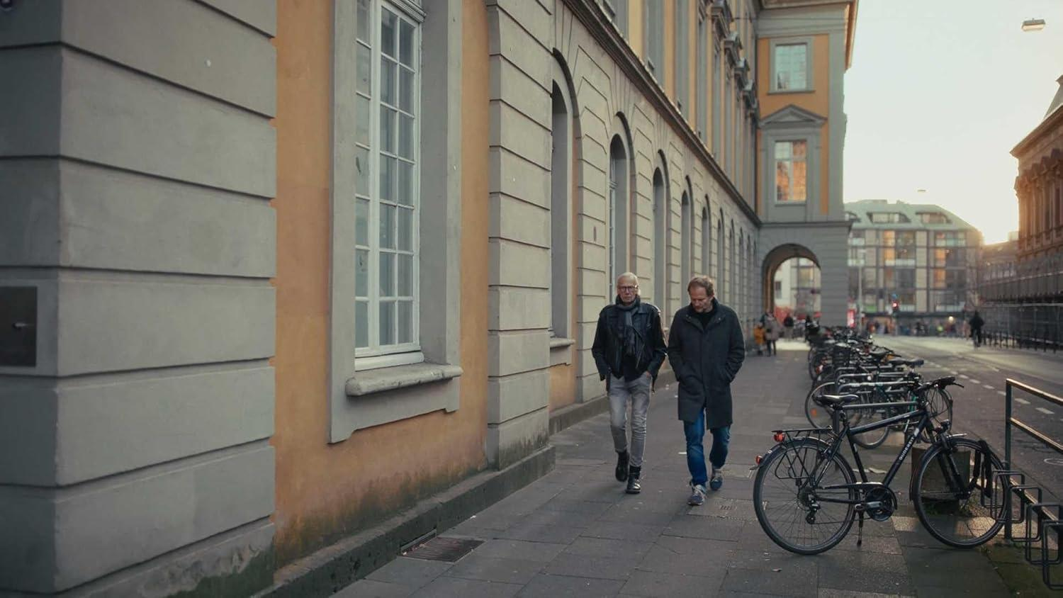 Zwei Männer gehen auf einem Bürgersteig neben einem Gebäude. Fahrräder sind am Straßenrand geparkt. Gebäude und Bögen sind im Hintergrund.