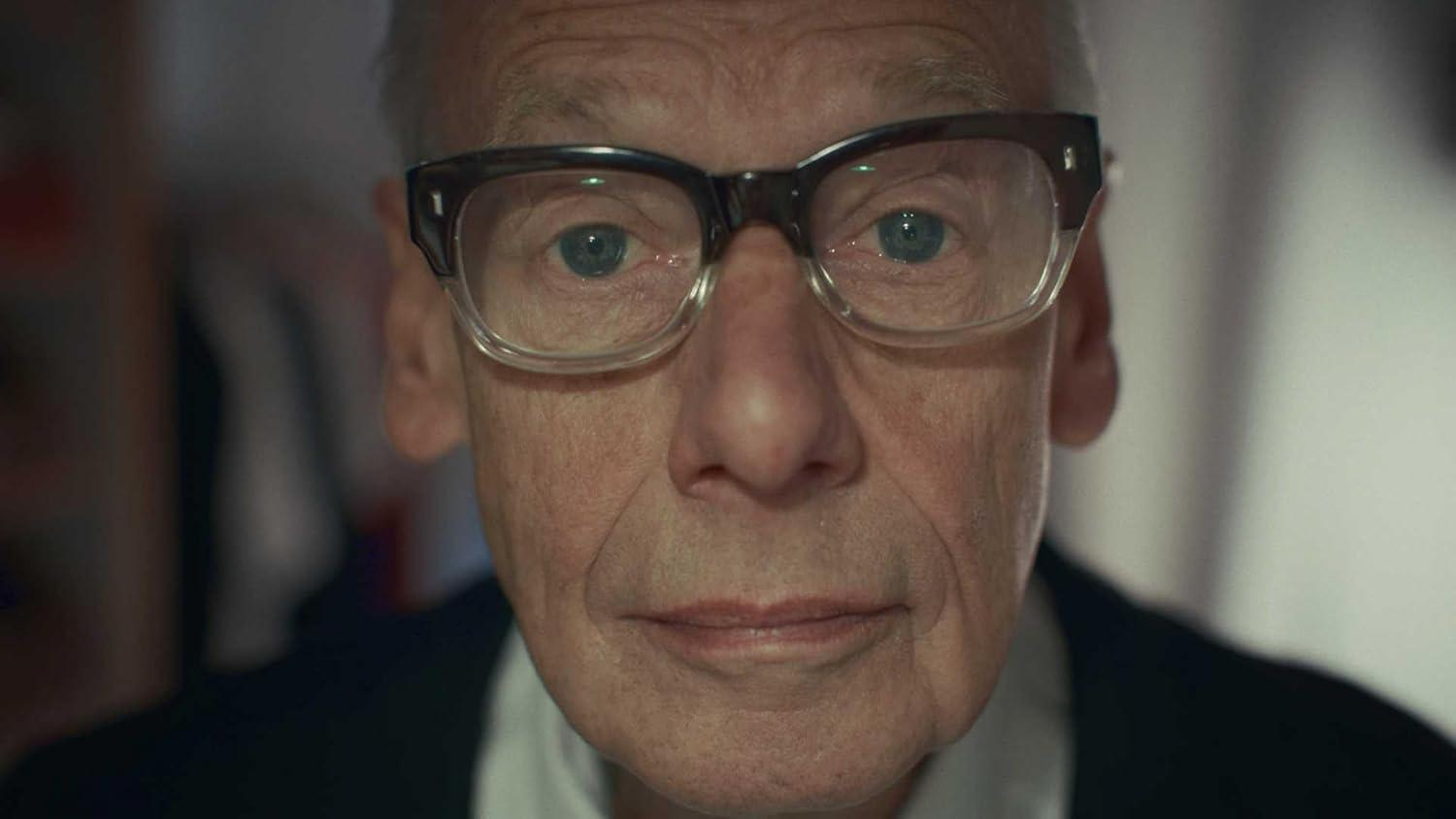 Nahaufnahme eines älteren Mannes mit Brille. Er hat einen hellen Teint, blaue Augen und einen neutralen Gesichtsausdruck. Der Hintergrund ist unscharf.