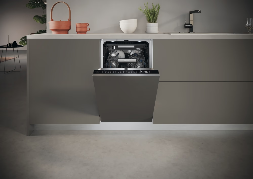 Nyitott mosogatógép modern konyhában. Belül edények. Teáskanna és csészék a pulton. Szürke szekrények és padló.