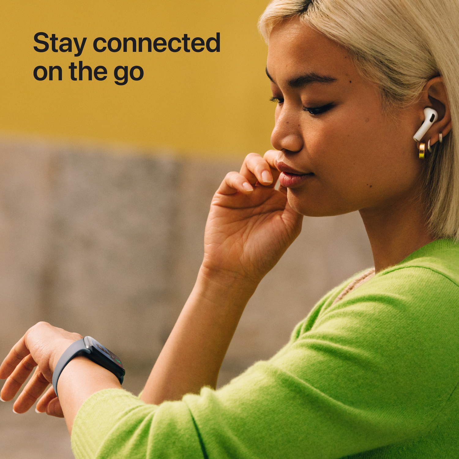 Vrouw met oortelefoons en smartwatch, kijkt naar het horloge. Tekst: Onderweg verbonden blijven. Groene trui, gele achtergrond.