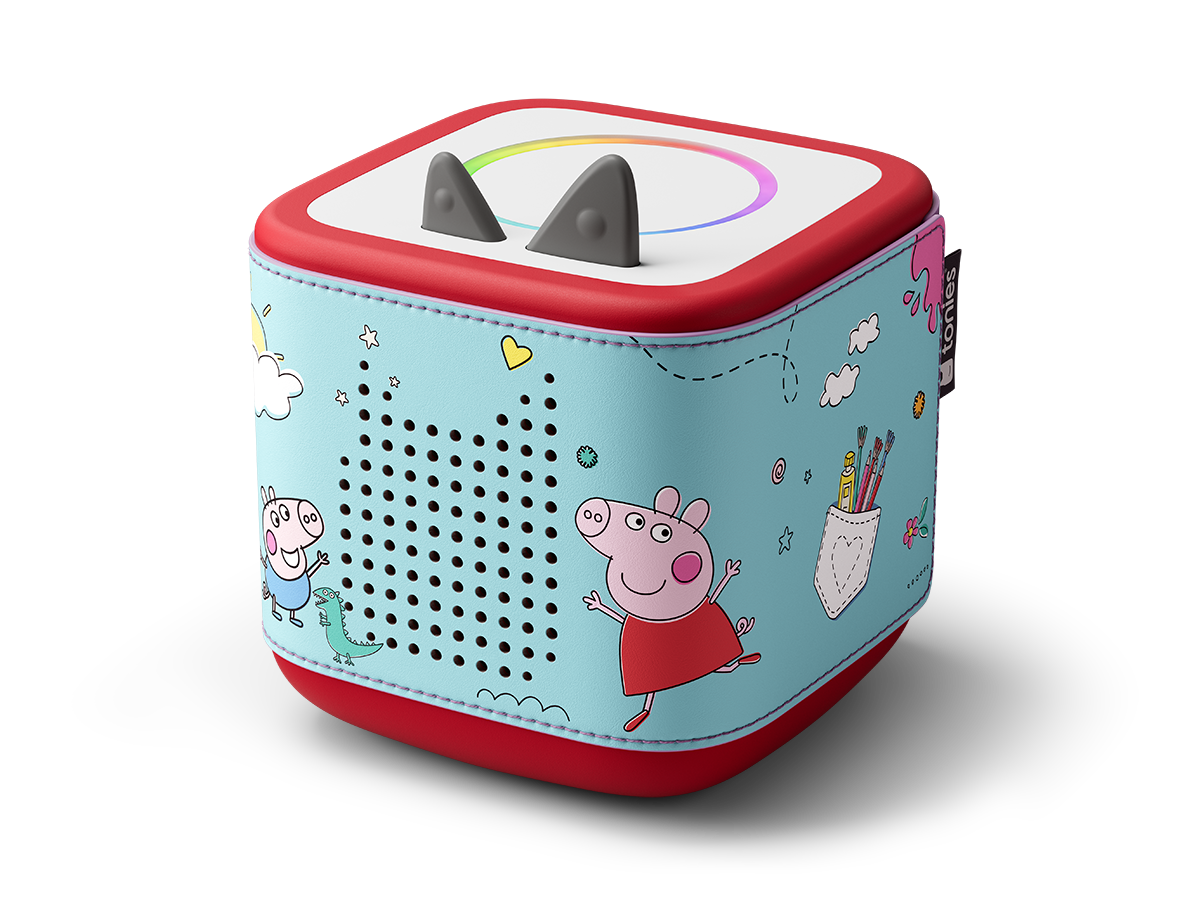 Un jouet en forme de cube avec des illustrations de Peppa Pig. Il a une base et un dessus rouges, et un corps bleu clair. Comprend des oreilles et un haut-parleur.