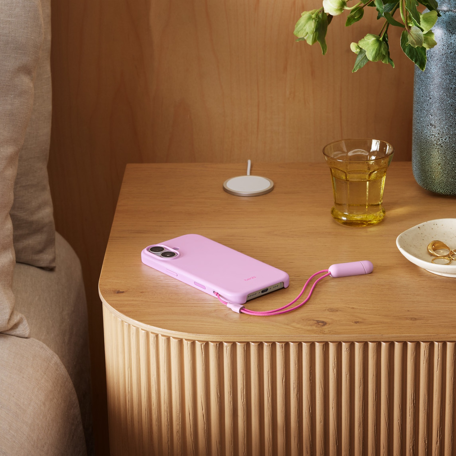 Een nachtkastje met telefoon, oplader, glas en vaas. De telefoon is roze, met een roze oplaadkabel. De achtergrond is van hout.