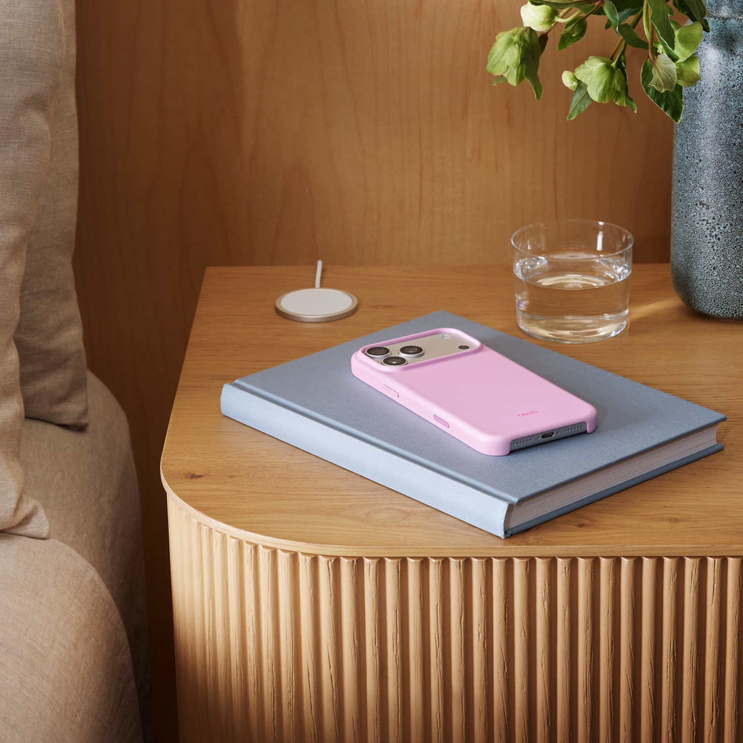 Un étui de téléphone rose est posé sur un livre gris sur une table de chevet en bois. Un verre d'eau et un chargeur blanc sont également présents.