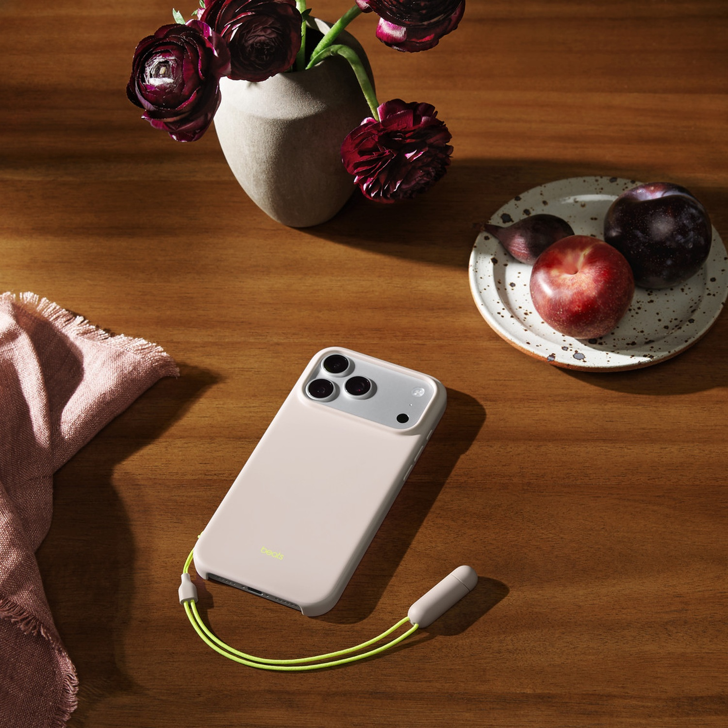 Roze telefoonhoesje met een limoengroene band op een houten tafel. Er zijn ook bloemen, pruimen en een roze servet aanwezig.