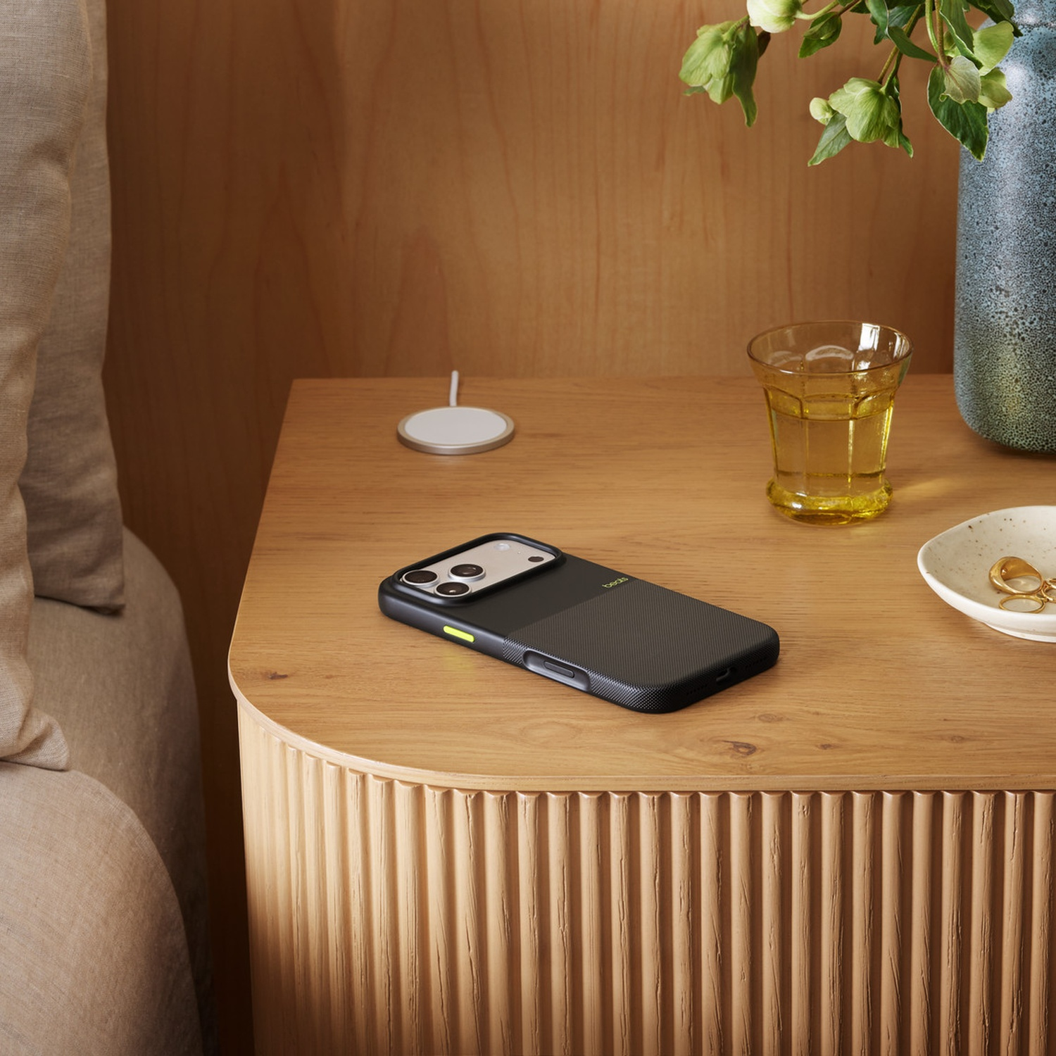 Een nachtkastje met een telefoon in een zwarte hoes, een glas vloeistof, een vaas met bloemen en een oplaadpad.