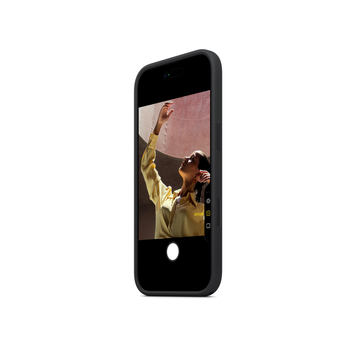 A smartphone displays a woman in a yellow shirt. She's raising her arm, with a pink wall background. The phone is black.