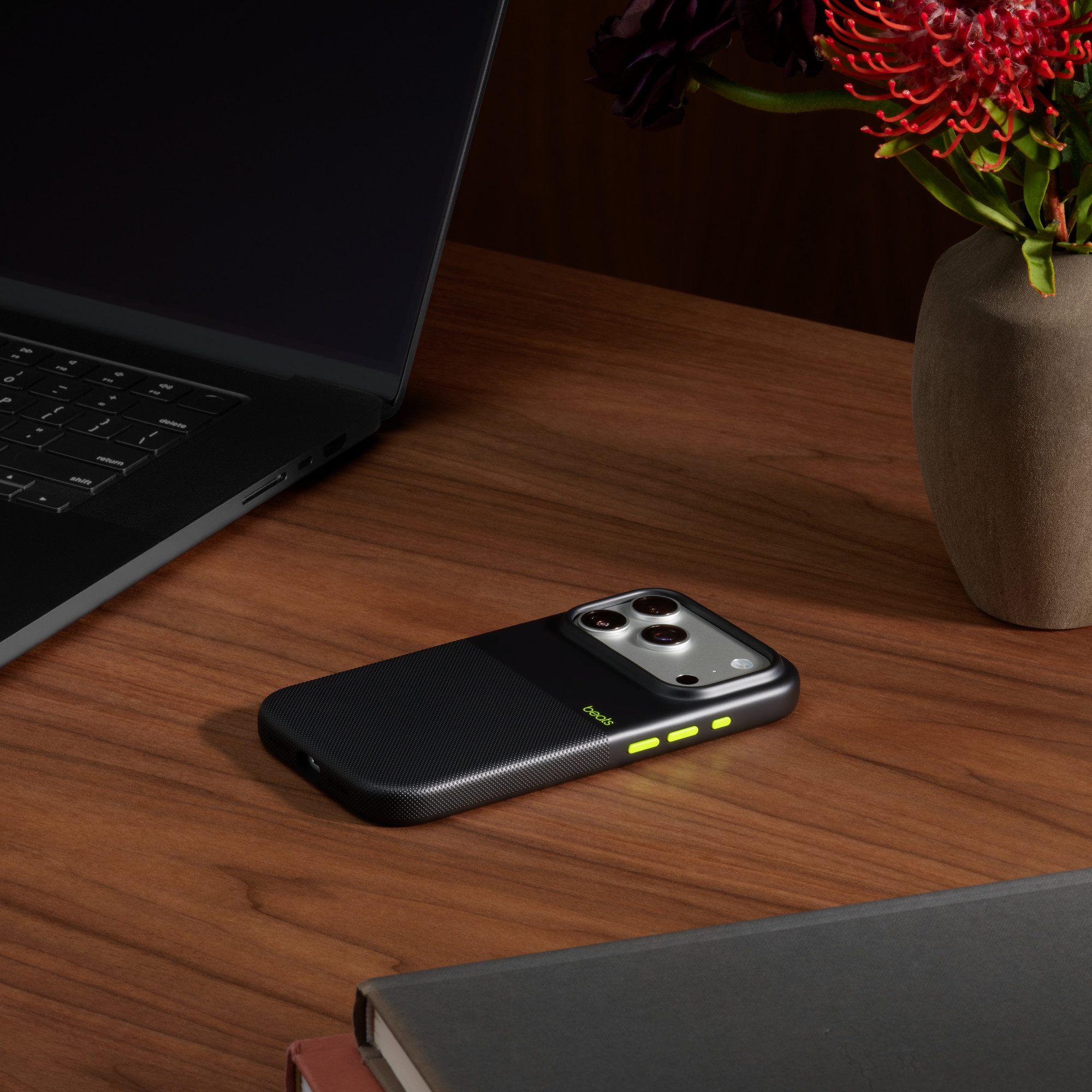 Czarny telefon z zielonym etui leży na drewnianym biurku obok laptopa, kwiatów i książek.