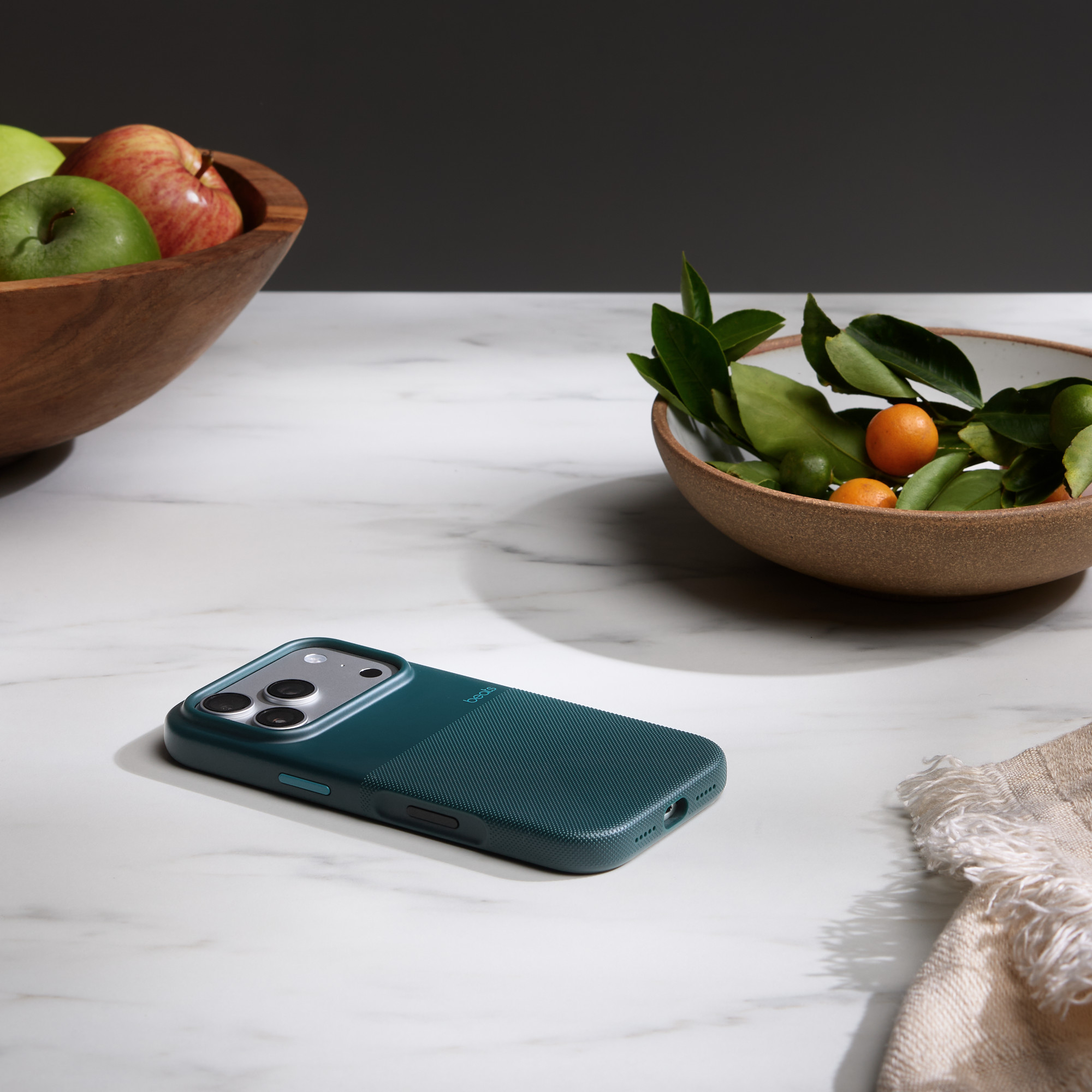 Teal etui na telefon leży na marmurowej powierzchni, z miseczkami owoców i zielenią. Miękkie światło i cień.