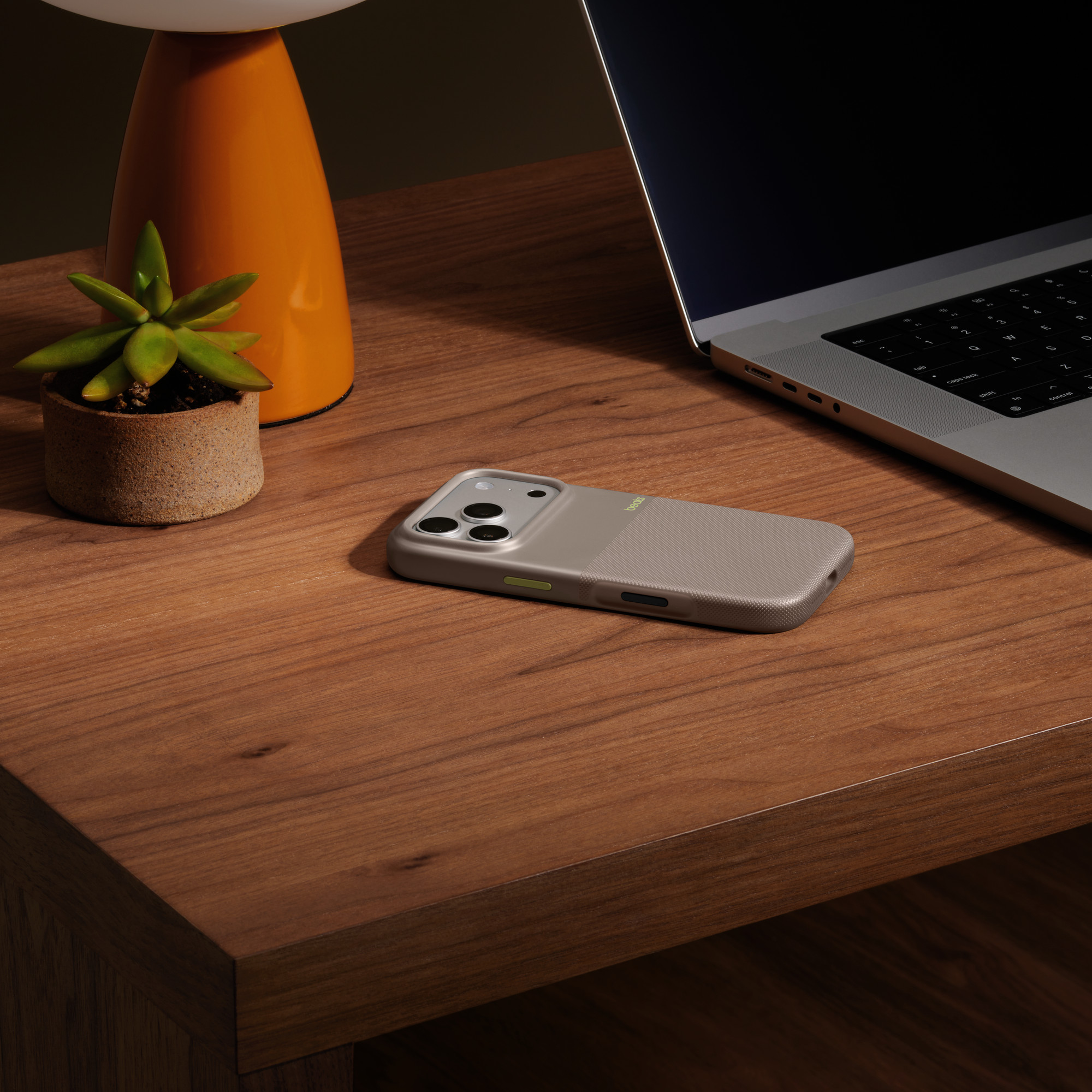 Etui na iPhone'a, laptop, roślina i lampa na drewnianym stole. Etui jest szare z zielonym akcentem.