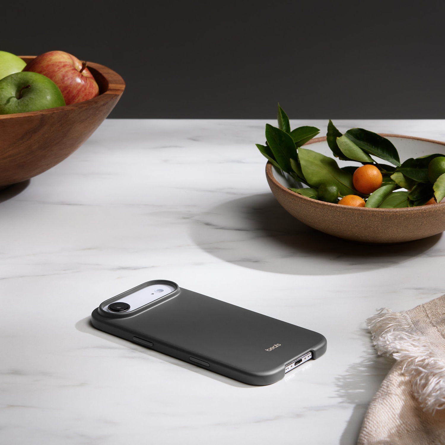 Een grijze telefoonhoes ligt op een marmeren oppervlak. Schalen met appels en citrusvruchten in de buurt, rechts een neutrale doek.