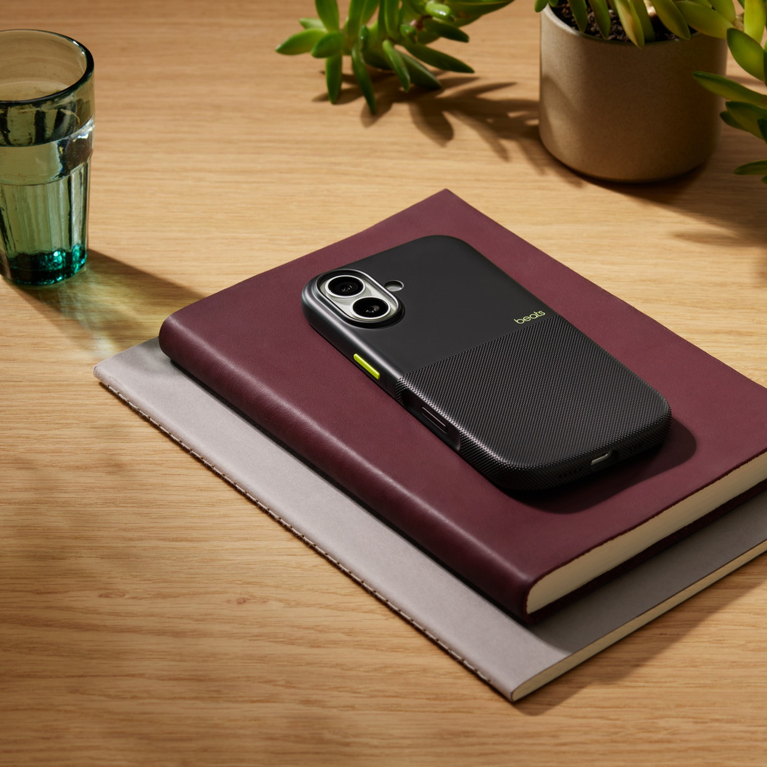 Un étui de téléphone noir sur un cahier bordeaux, avec un verre d'eau et une plante sur une table en bois. L'étui porte le logo 'beats'.