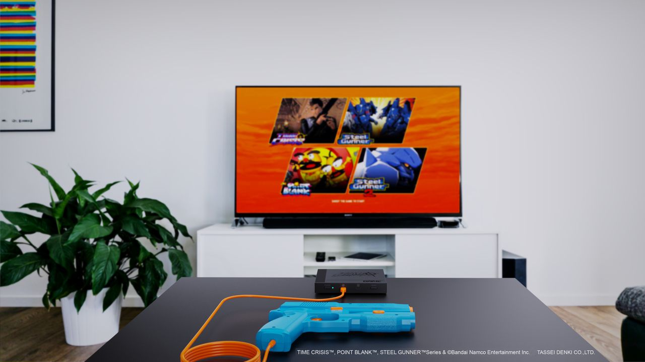 Una pistola azul en una mesa está conectada a una consola negra. Un televisor muestra opciones de juego. Una planta y un arte colorido enmarcado a la izquierda.