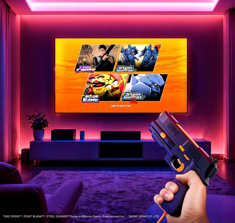 Una persona sostiene una pistola de juguete en una habitación con una pantalla grande que muestra opciones de juego. La habitación está iluminada con tonos púrpuras.