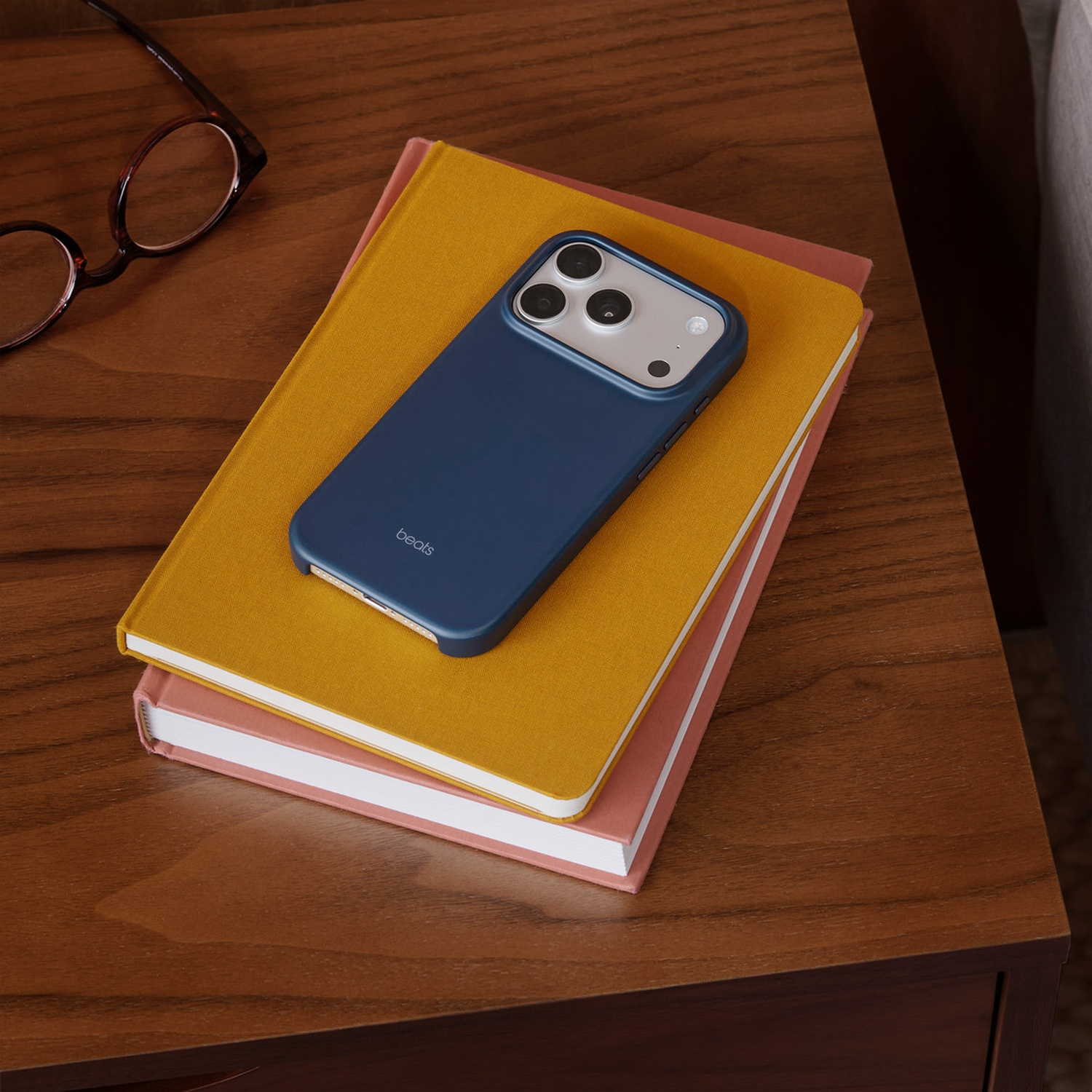 Un étui de téléphone bleu est posé sur deux livres, jaunes et roses, sur une surface en bois. Des lunettes sont à côté.