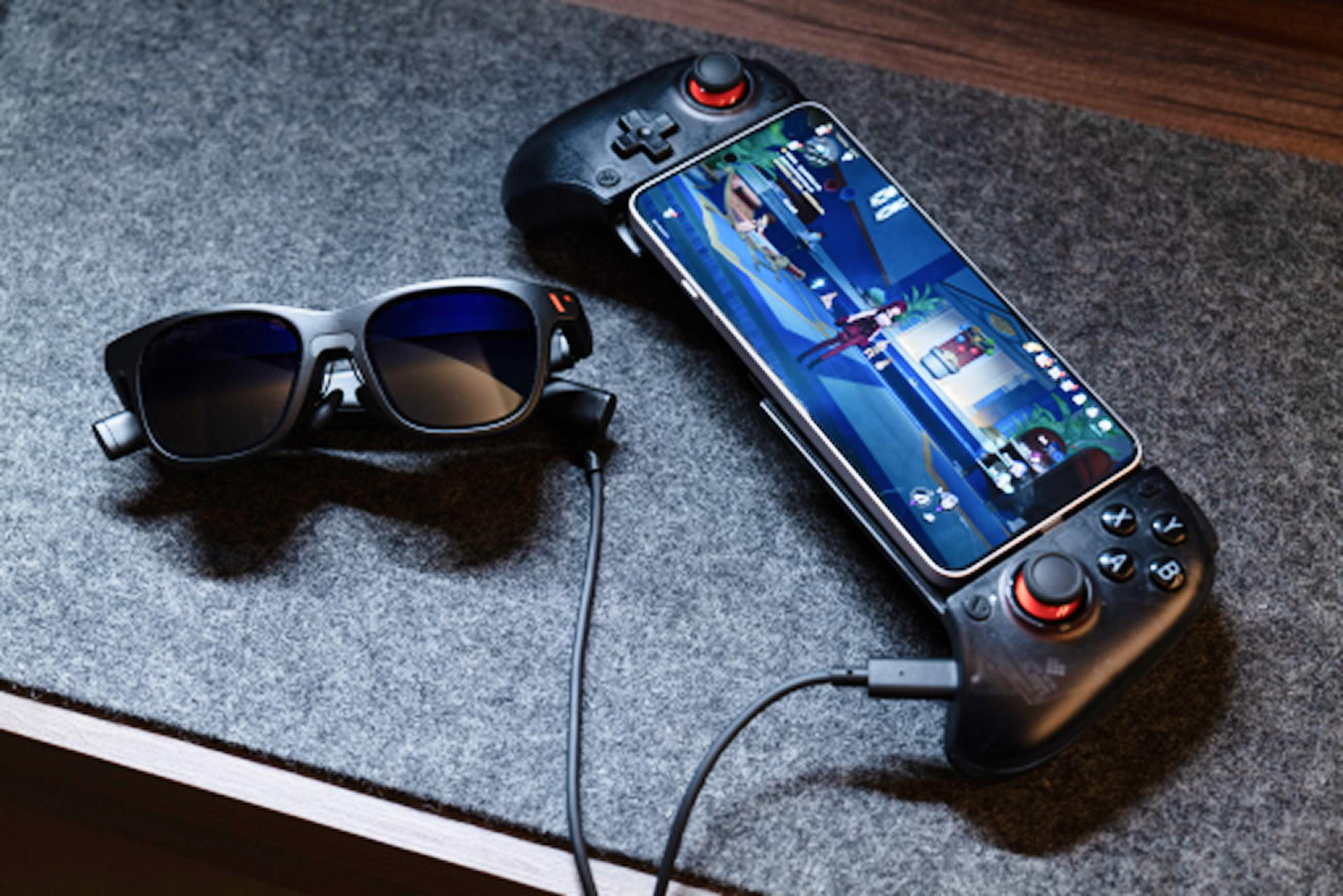 Eine schwarze Brille, ein Smartphone mit Spiel und ein Gamepad liegen auf einer grauen Oberfläche. Ein Kabel verbindet die Brille.