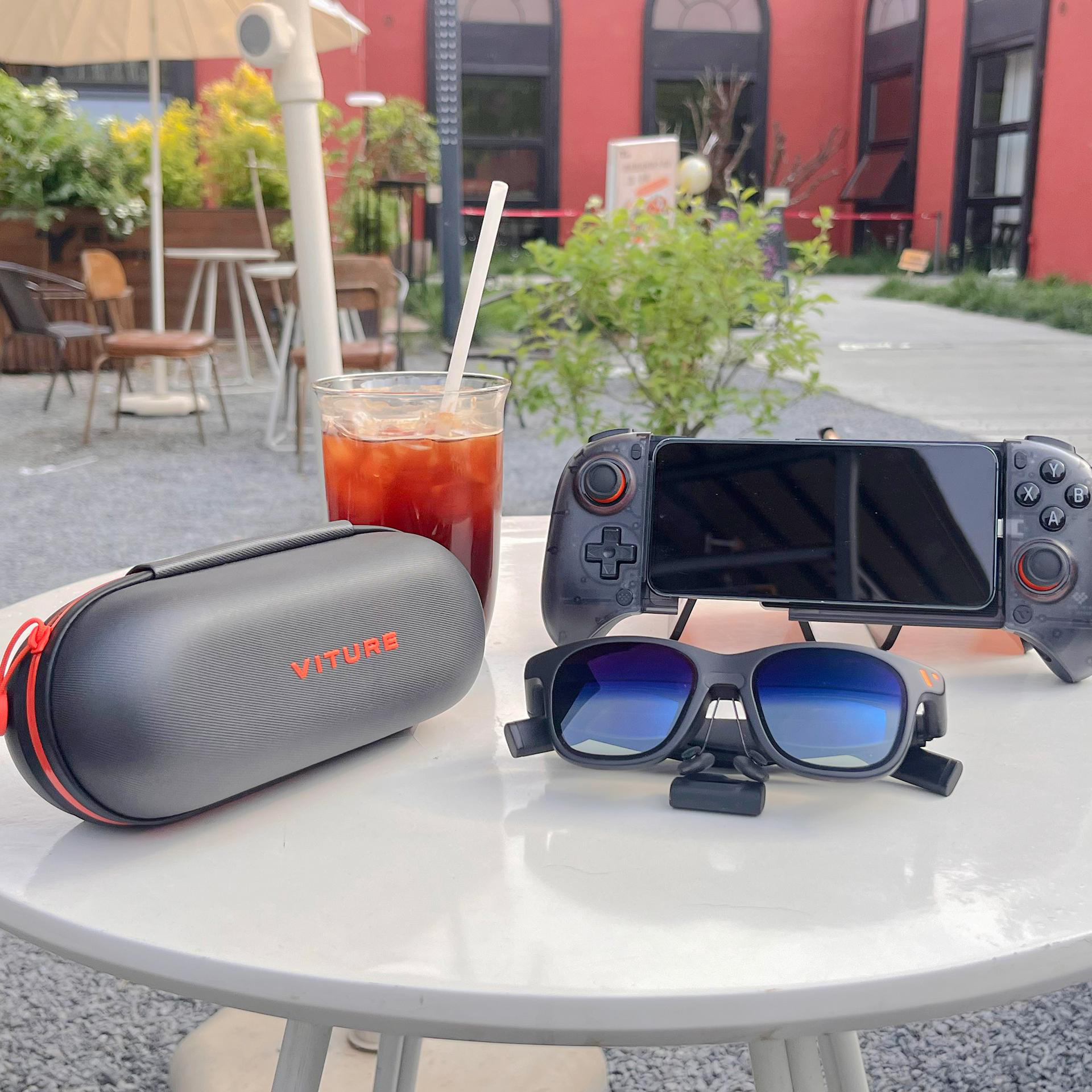 Tisch mit Brille, Controller, Getränk und Etui. Rotes Gebäude und Sitzgelegenheiten im Freien im Hintergrund.