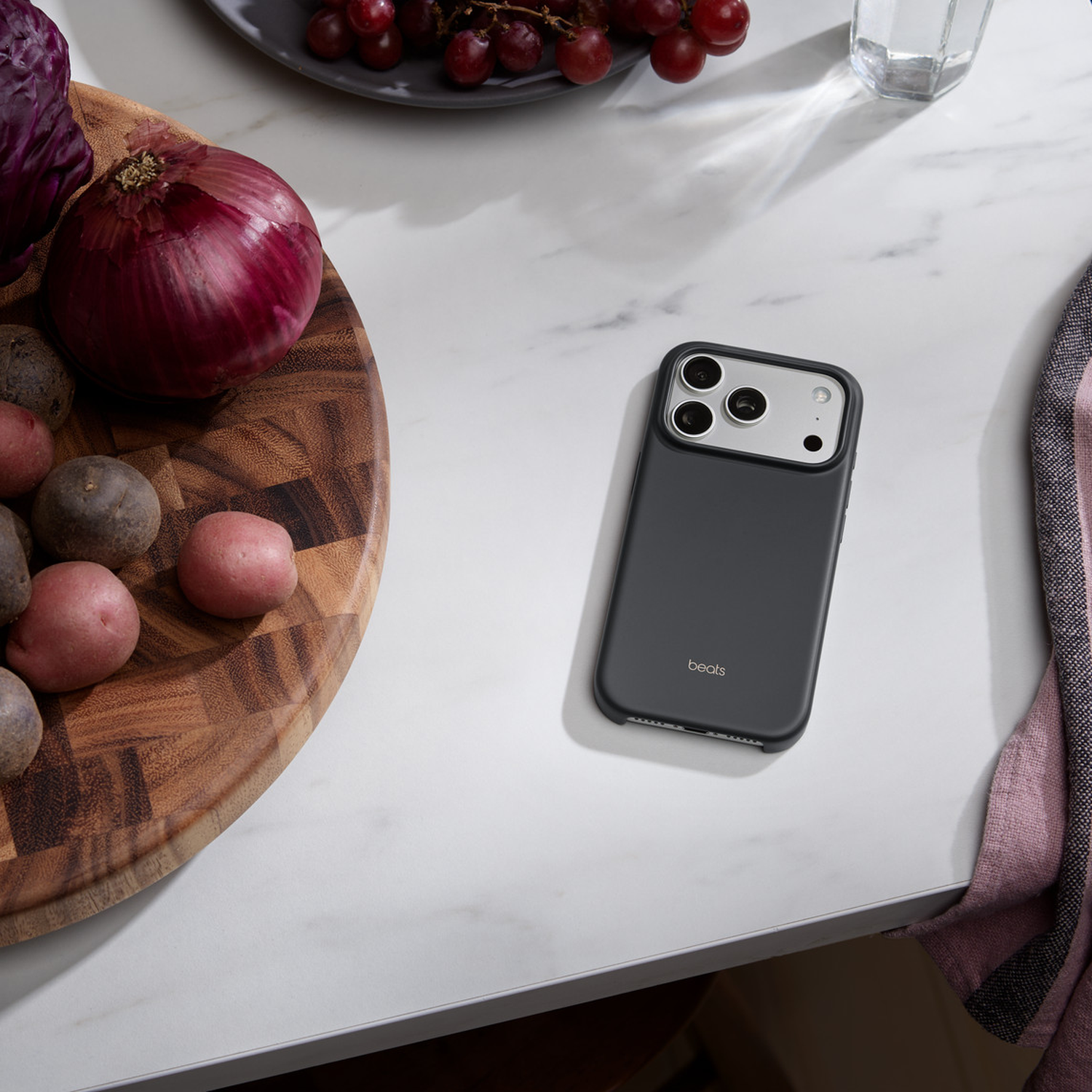 Een telefoon, groenten en druiven liggen op een marmeren oppervlak. Een snijplank bevat aardappelen en een ui. Een glas en een handdoek zijn ook aanwezig.