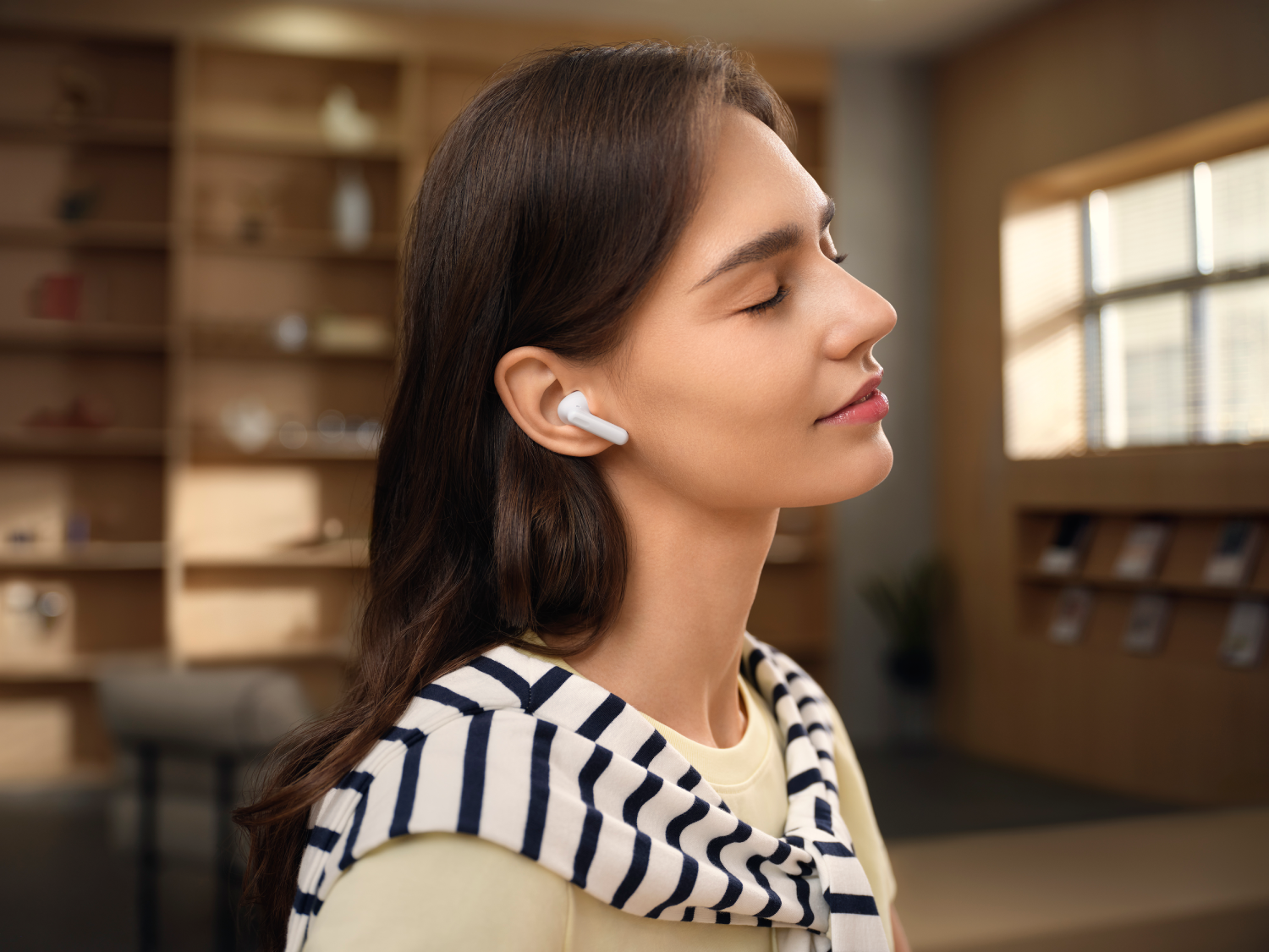 Una mujer con auriculares cierra los ojos. Lleva un chal a rayas y una camisa amarilla. Estanterías y una ventana al fondo.