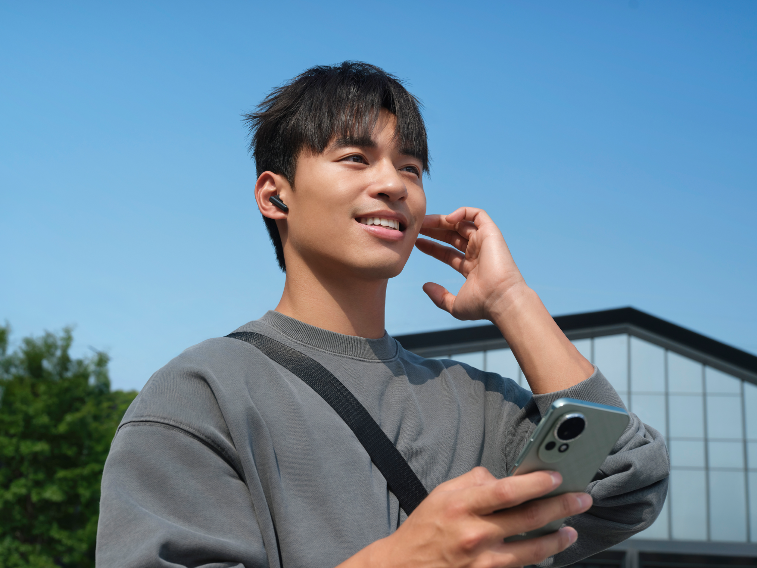 Un hombre sonriente con auriculares, sosteniendo un teléfono y una bandolera. Edificio moderno y cielo azul de fondo.