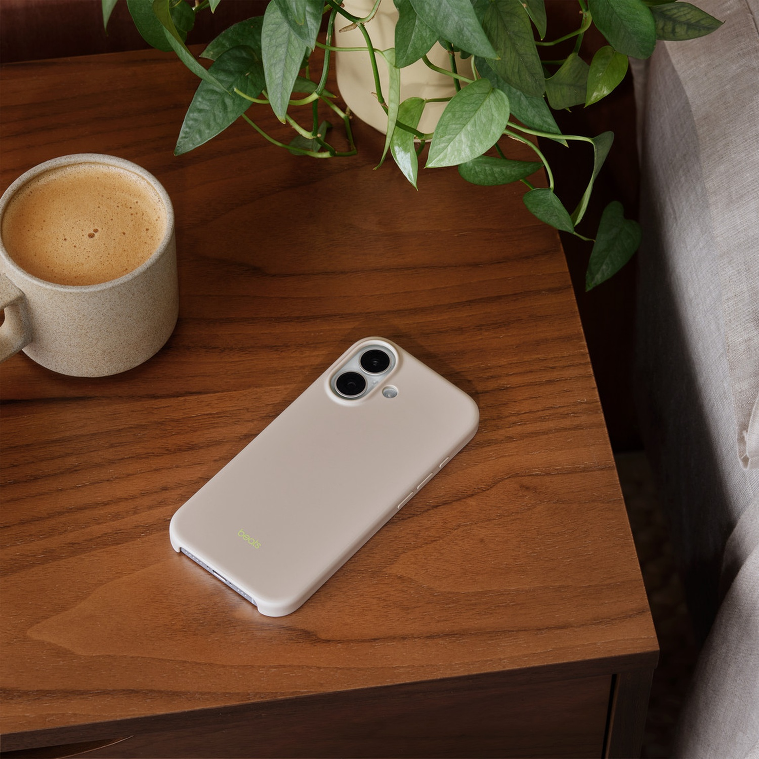 Een beige telefoon op een houten tafel naast een koffiemok en een plant. Een bed staat op de achtergrond.