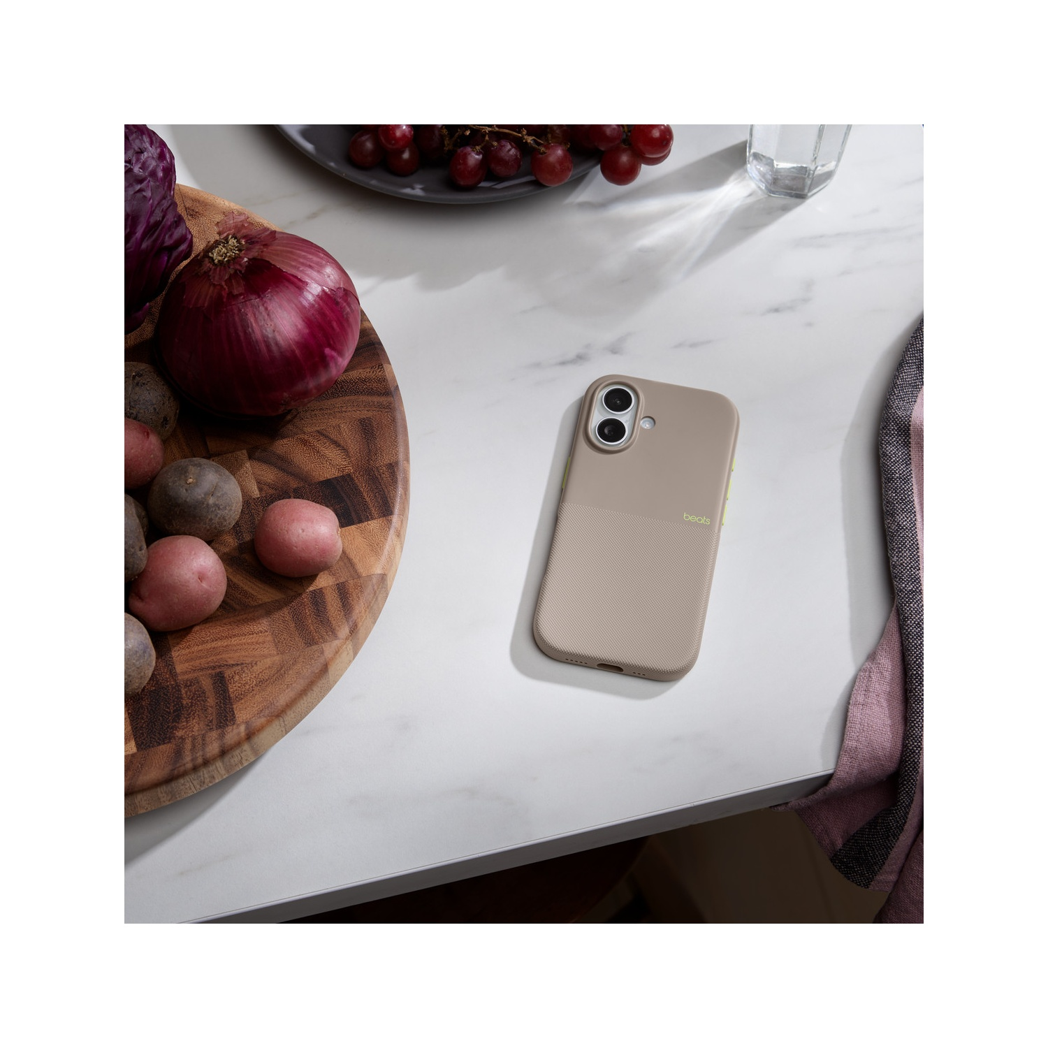 Une coque de téléphone, des légumes, des raisins et un verre d'eau sur une surface blanche. Une planche à découper en bois avec des pommes de terre et un oignon est visible.