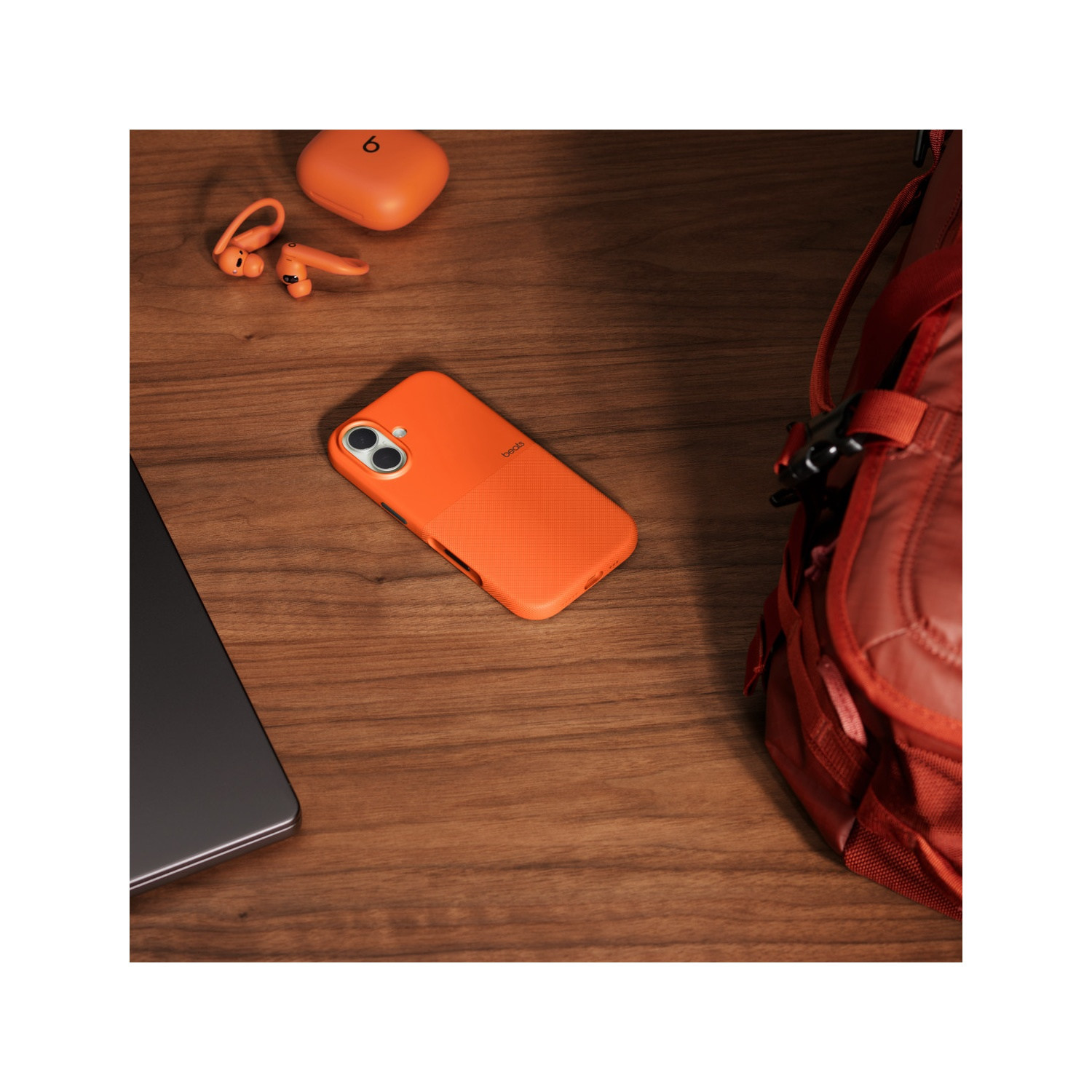 Oranje telefoonhoesje, oordopjes en oplaadcase op een houten tafel naast een rugzak en een laptop.