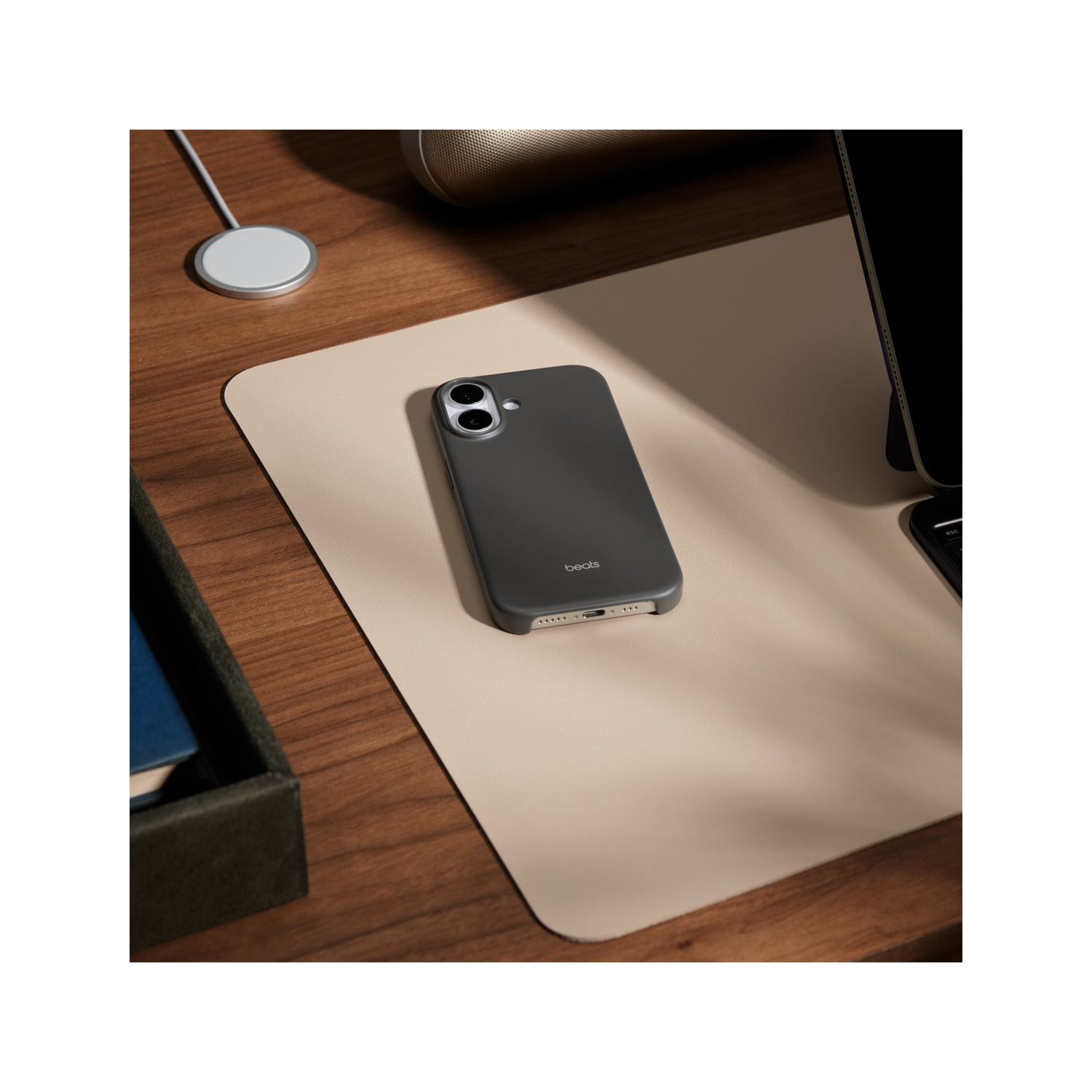 Un étui de téléphone Beats gris foncé sur un tapis de bureau beige. Un chargeur blanc et un haut-parleur en arrière-plan. Table en bois.